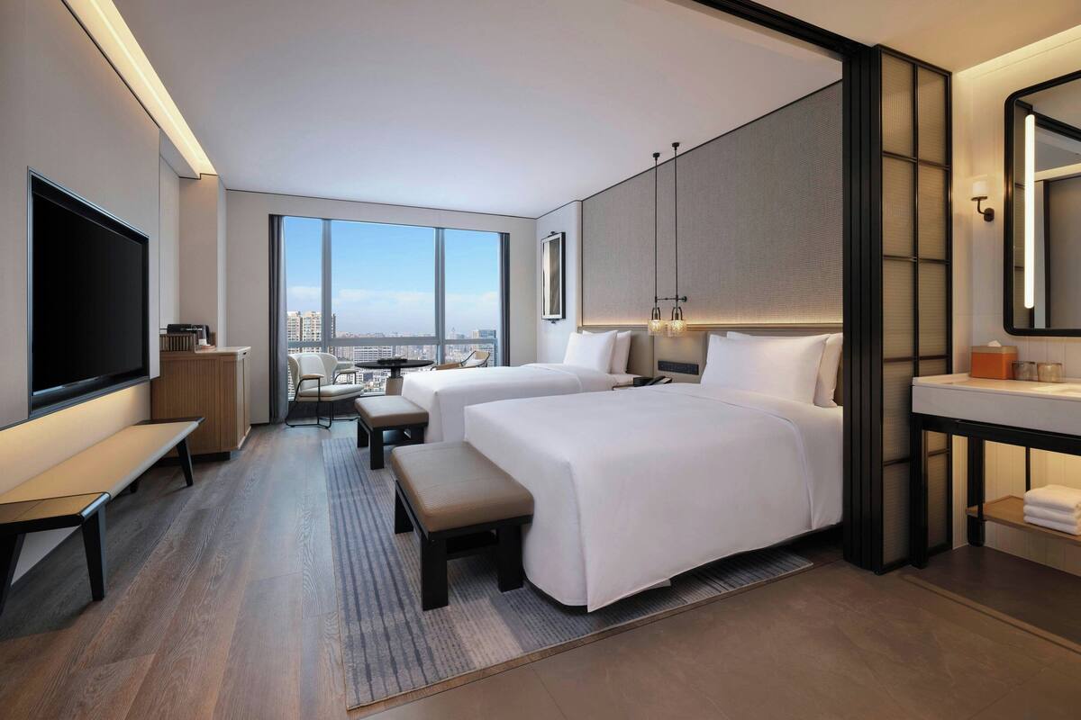 Quarto de hotel moderno com duas camas, uma televisão grande, uma escrivaninha e vista para a cidade.