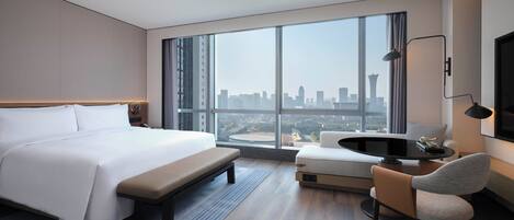 Un cuarto de hotel moderno con una cama grande, un espacio de descanso con una mesa y vistas a la silueta urbana a través de amplias ventanas.