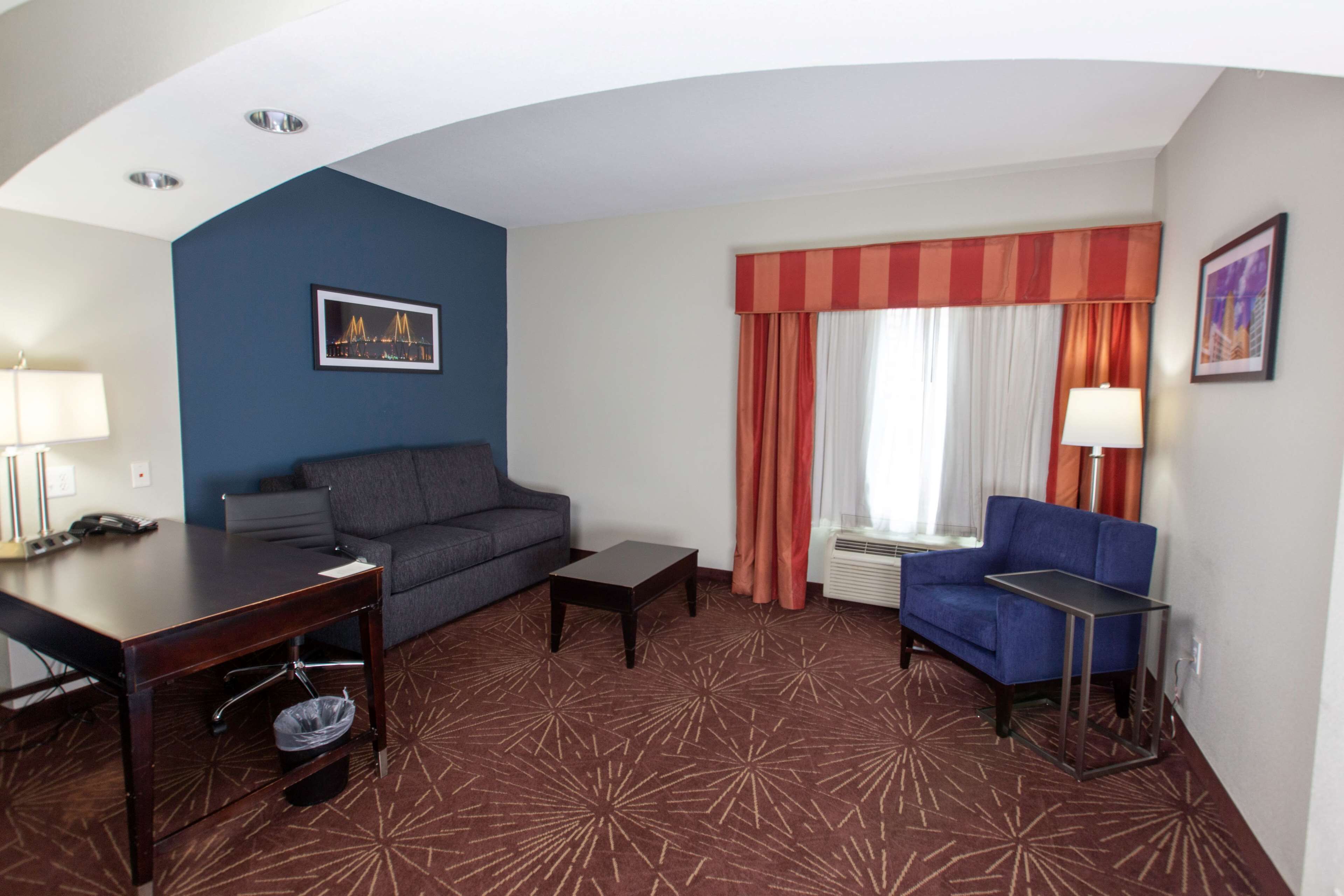 Hotelkamer met een blauwe accentmuur, een grijze bank, een blauwe fauteuil, een kleine tafel, een bureau met telefoon en een raam met gordijnen.