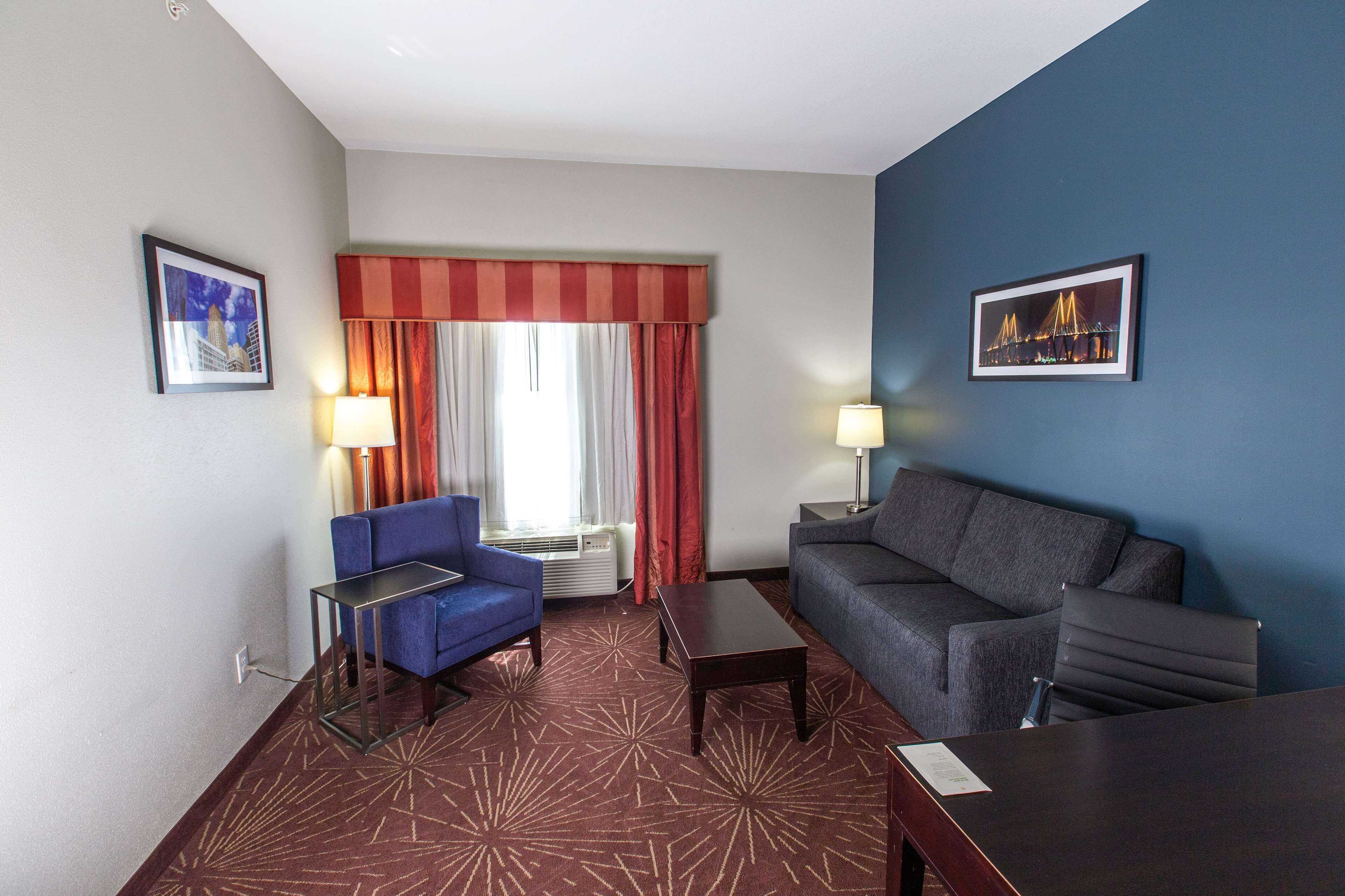 Une chambre d’hôtel avec un canapé bleu et gris, une petite table, une lampe et une fenêtre avec des rideaux rouges.