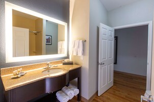 Un baño con un espejo grande, un lavabo y un inodoro. Hay toallas colgadas en la pared y una puerta que da a otra habitación.