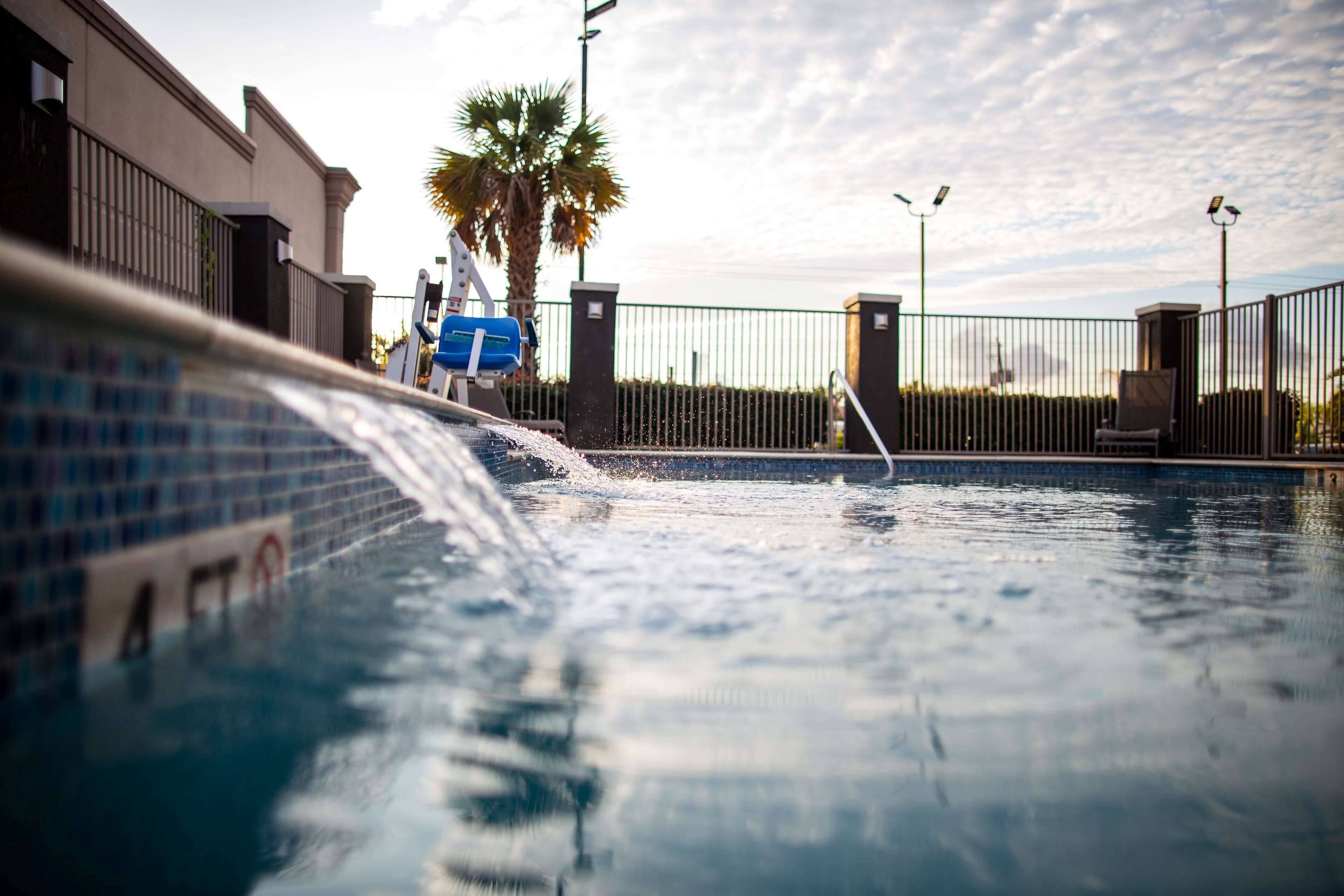 A swimming pool with a water feature, surrounded by a fence, palm trees, and a building with a tiled facade.