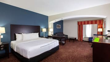 Habitación de hotel con una cama grande, un escritorio, una silla y una mesita. Hay una ventana con cortinas y un cuadro en la pared.