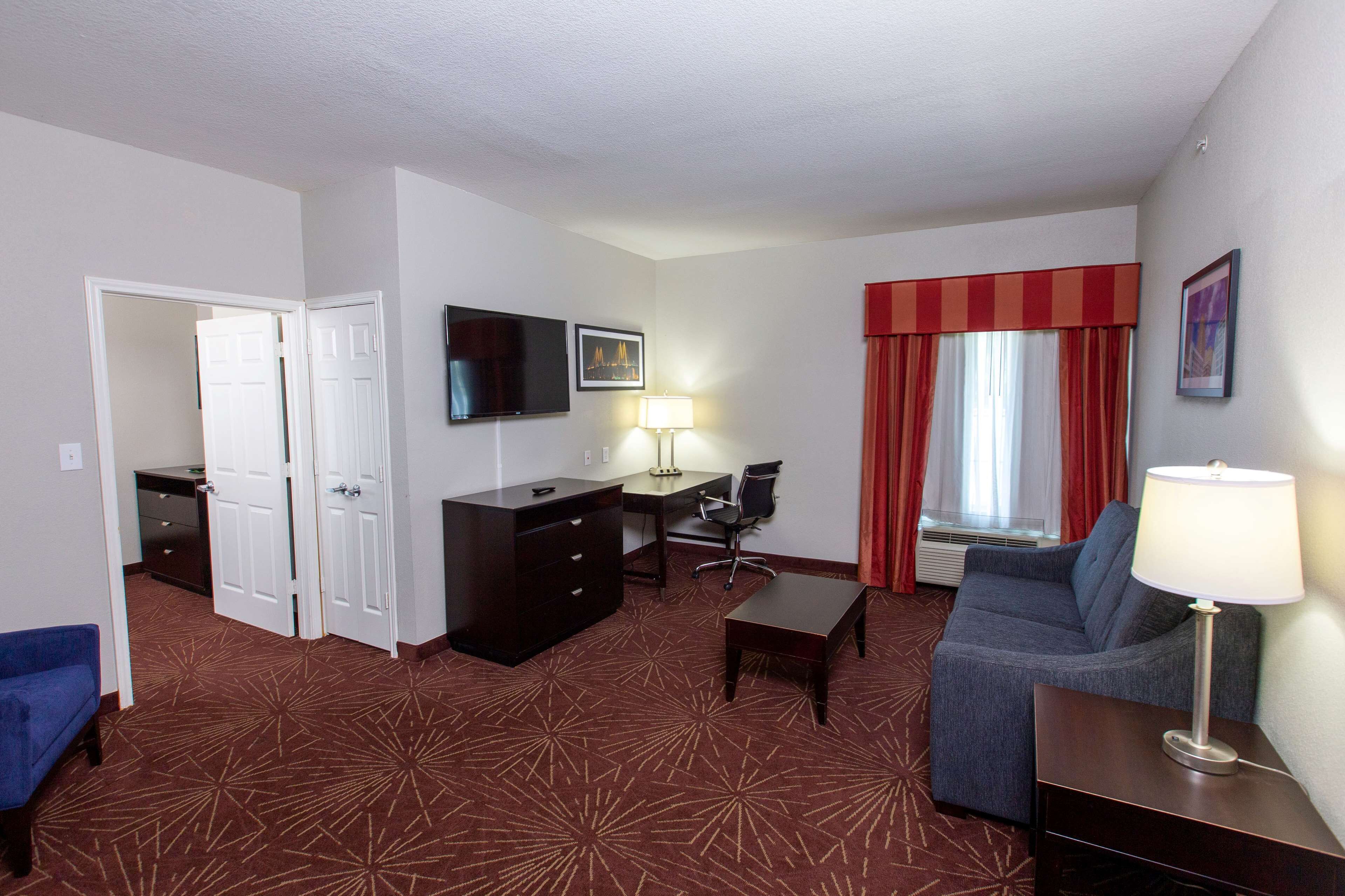 A hotel room with a red carpet, a sofa, a desk with a chair, a TV on the wall, and a window with red curtains.