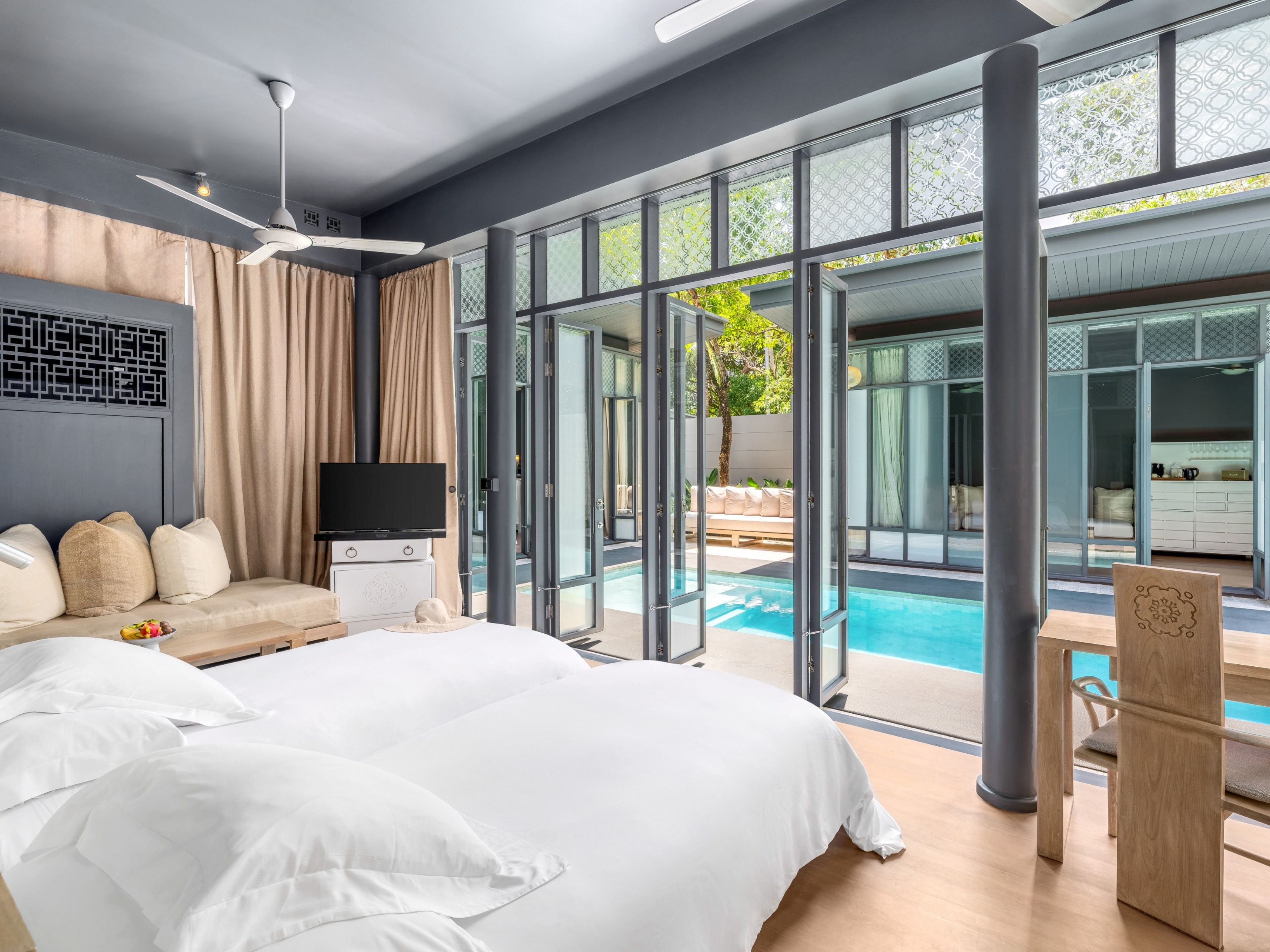 Habitación de hotel moderna con una cama grande, un televisor de pantalla plana, una zona de estar con sofá y cojines, y vistas a una piscina exterior.