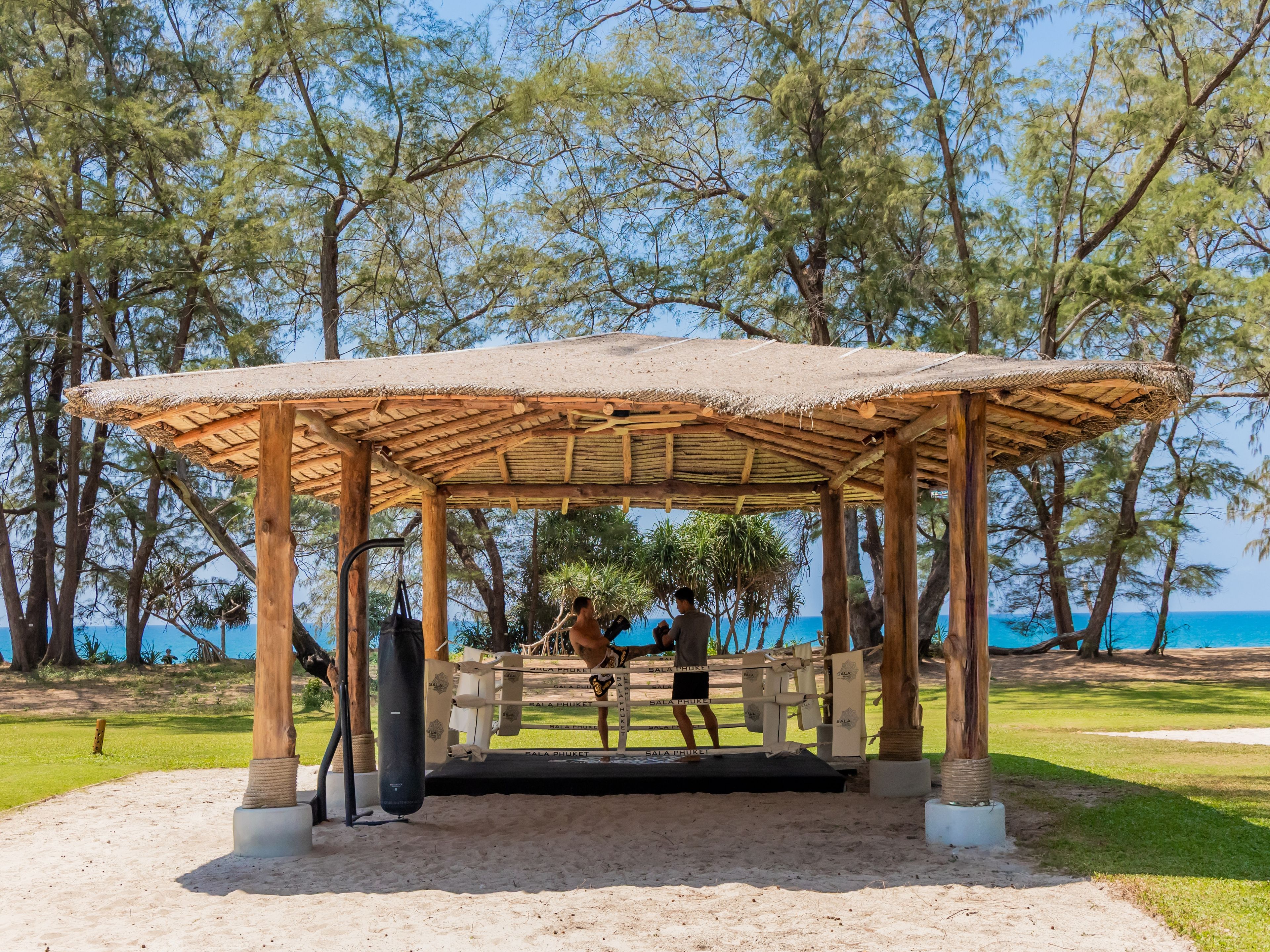 A wooden pavilion with a thatched roof, surrounded by trees and a clear view of the sea.