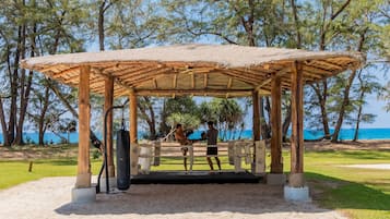 A wooden pavilion with a thatched roof, surrounded by trees and a clear view of the sea.