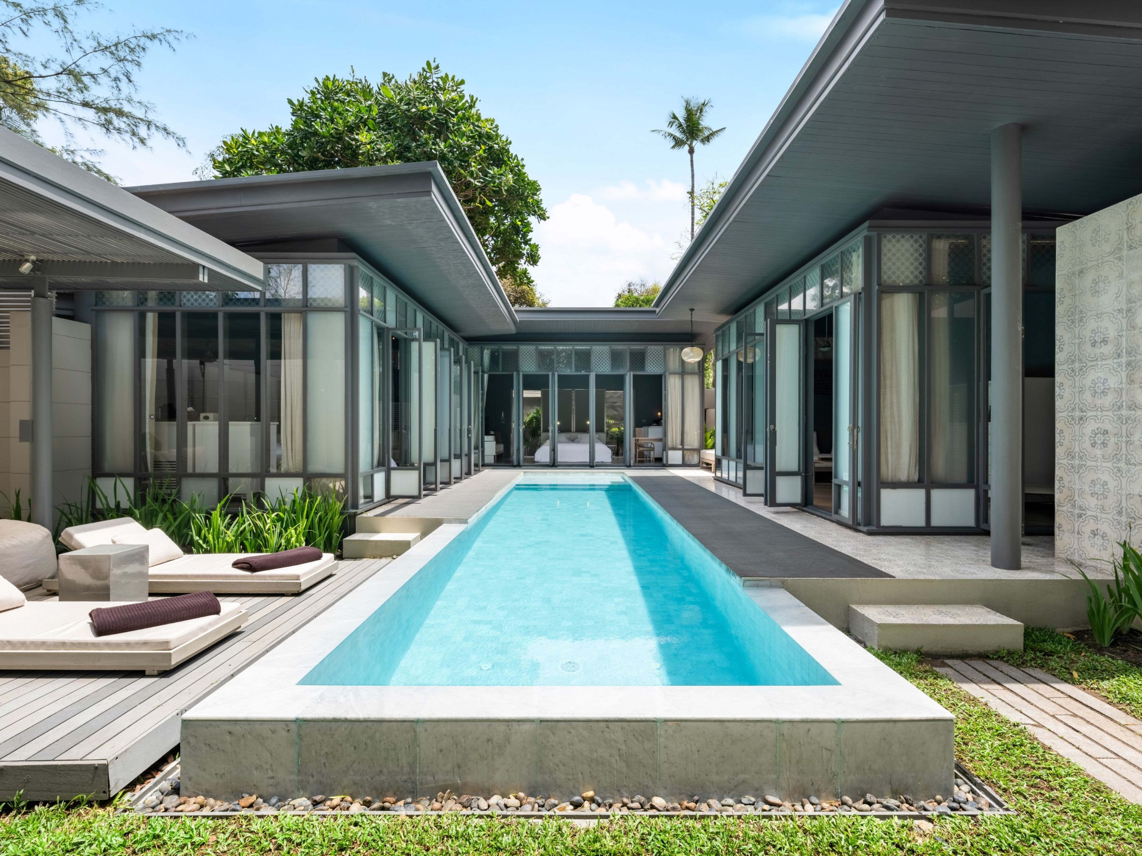 Un área de piscina moderna con una piscina rectangular, sillas de descanso y un edificio con cerramiento de vidrio.
