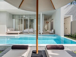 Un área de piscina moderna al aire libre con sillones, una cama y una sala con paredes de vidrio.