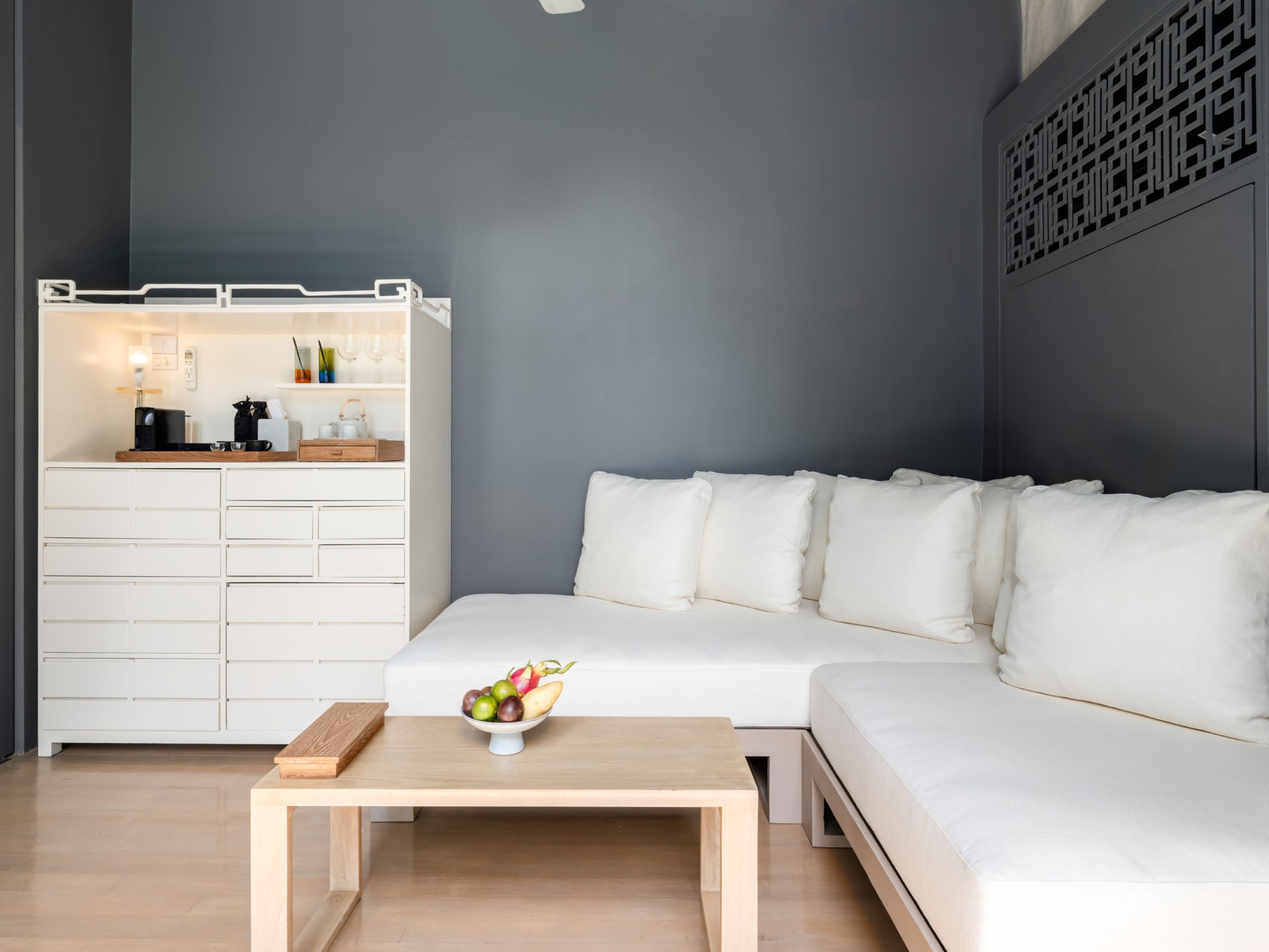 A modern living room with a white sofa, a wooden coffee table with fruit, and a white storage unit with kitchen appliances.