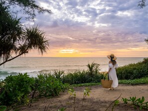 Une personne se tient sur une plage de sable, avec vue sur l’océan et un coucher de soleil.
