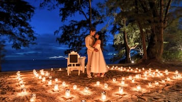 A beach setting with a couple standing by a table surrounded by candles, overlooking the sea.