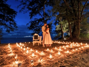 Escena de playa con una pareja de pie junto a una mesa rodeada de velas, con vistas al mar.