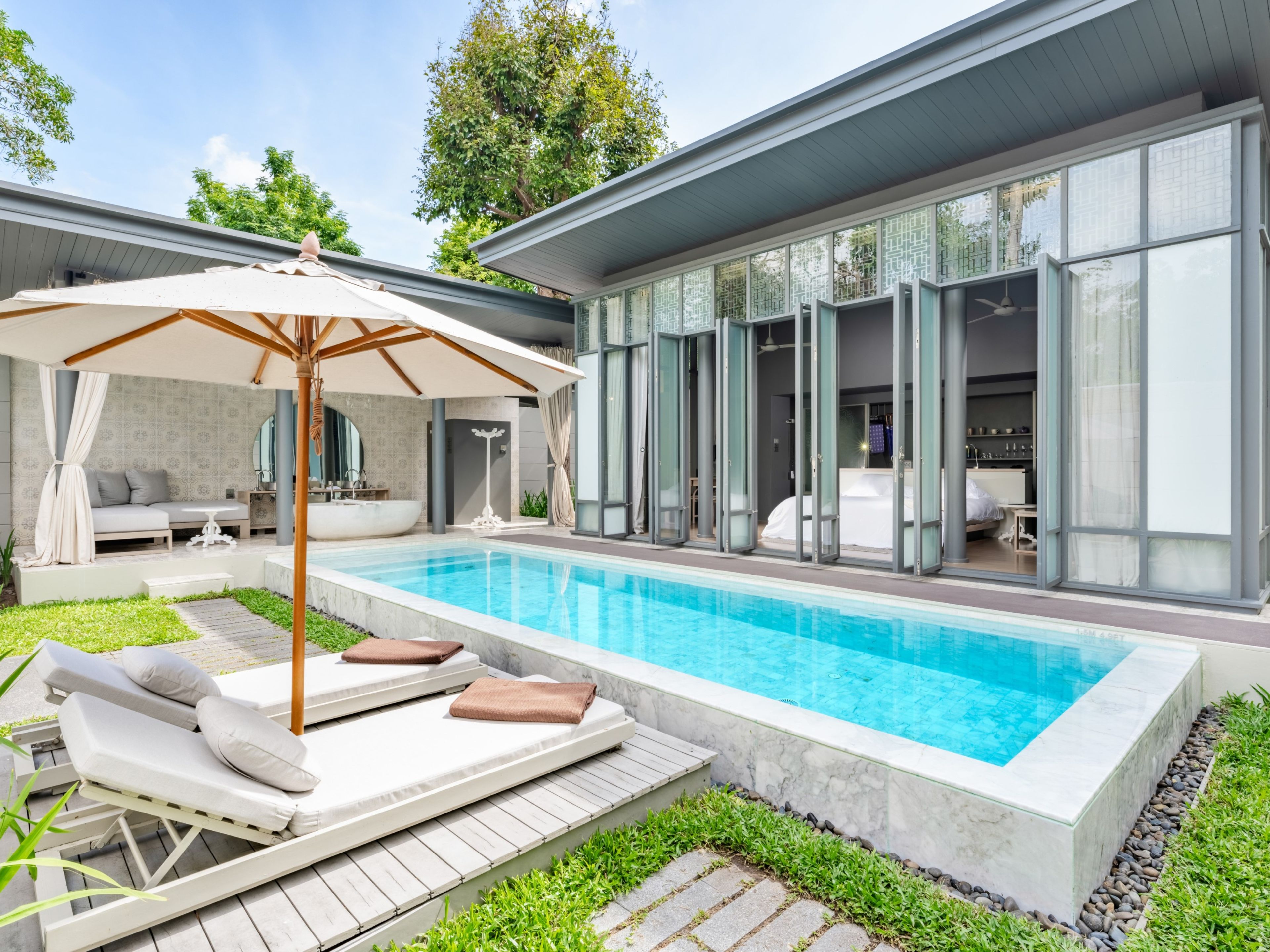 Un área moderna de piscina al aire libre con sillones de descanso y una sombrilla, rodeada por un jardín bien cuidado y un edificio contemporáneo con amplias ventanas.