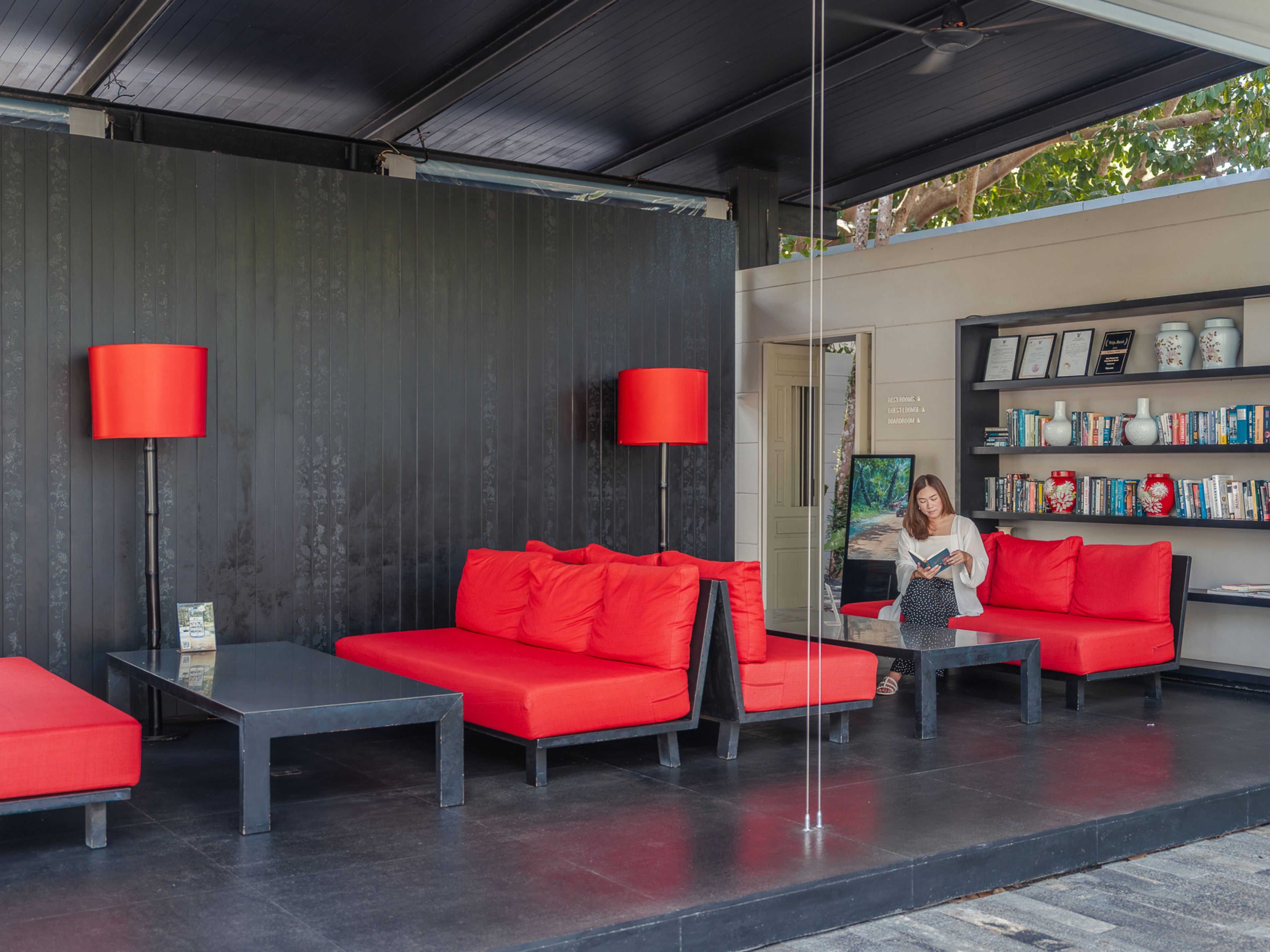 Un área de descanso moderna con sofás rojos y mesas negras, una estantería con libros y objetos decorativos, y una puerta que da a otra habitación.