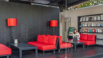 Un área de descanso moderna con sofás rojos y mesas negras, una estantería con libros y objetos decorativos, y una puerta que da a otra habitación.