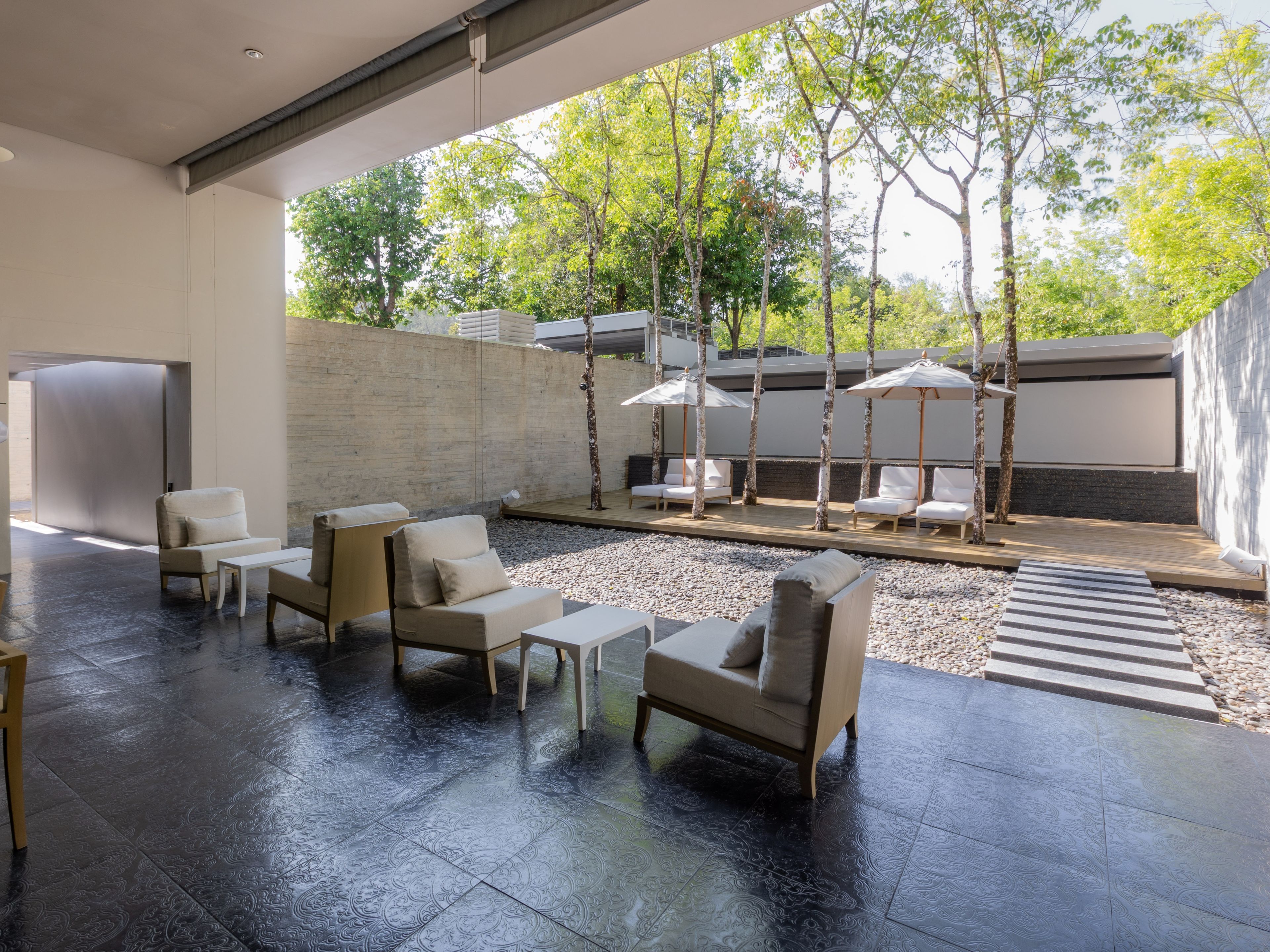 Un espacio exterior moderno con sillones de descanso y mesas, rodeado por un muro de piedra y árboles.