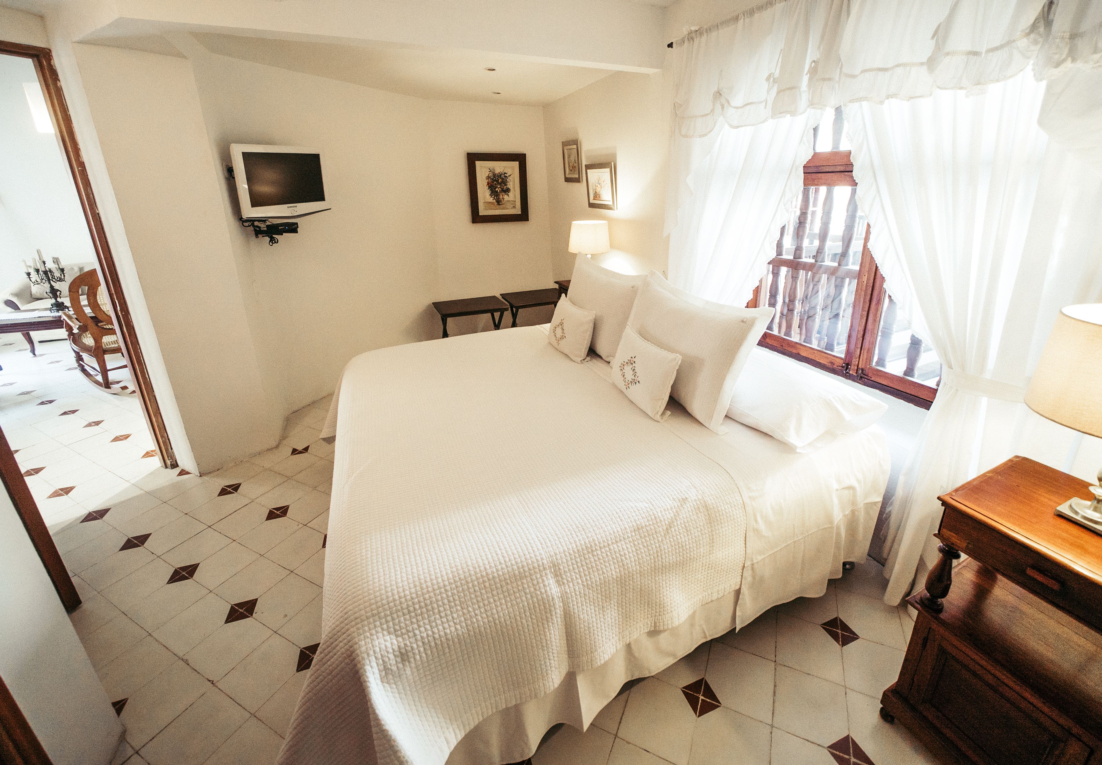 A neatly arranged bedroom with a large bed, bedside tables, a TV mounted on the wall, and a window with curtains.