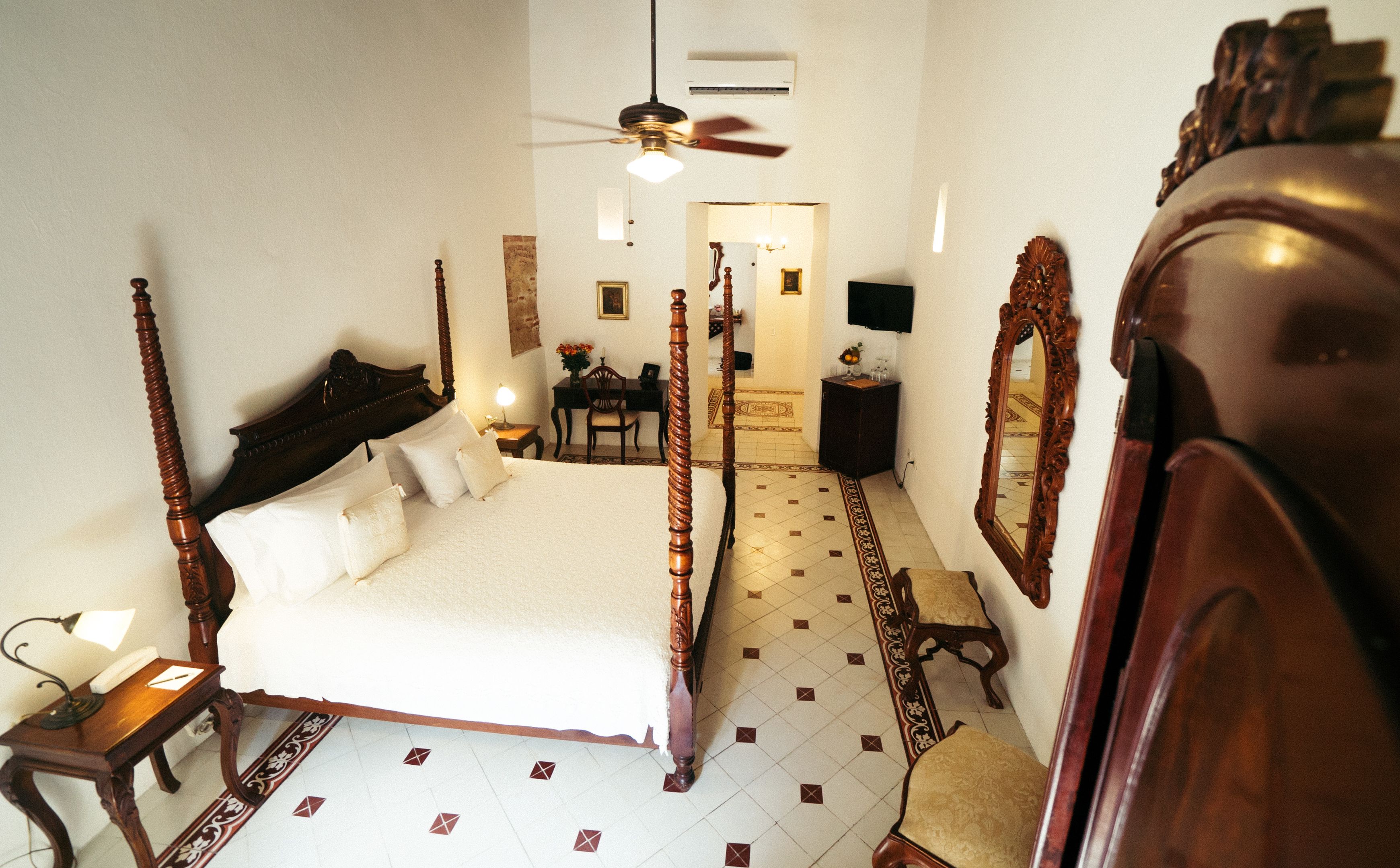 Una cama con dosel y tablero de madera, un escritorio con silla, un espejo y un ventilador de techo en una habitación de hotel.