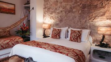 A bedroom with a brick wall, a hammock, a bed with patterned bedding, and a nightstand with a lamp.