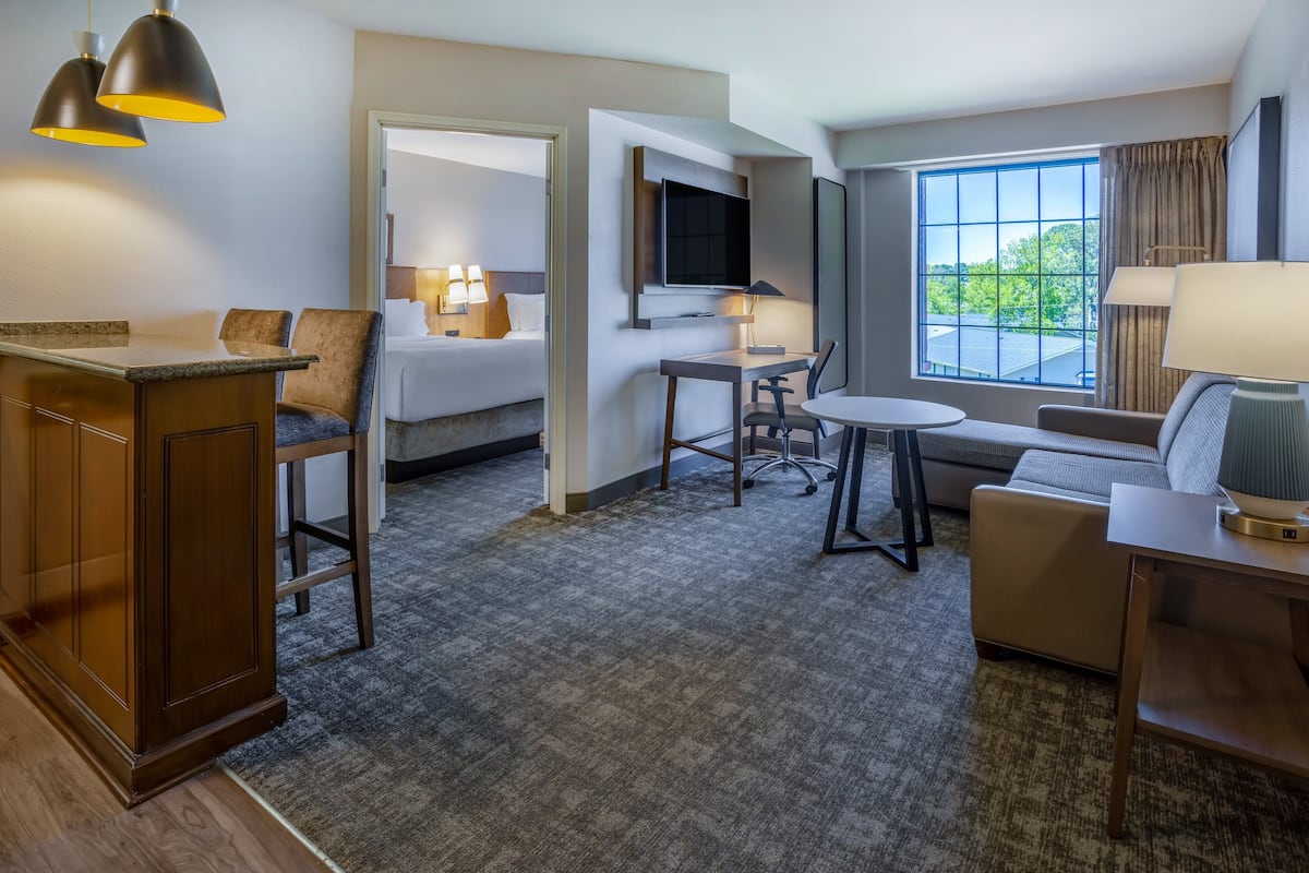 A hotel room with a bed, a desk, a chair, a sofa, a TV, and a window with a view of trees.