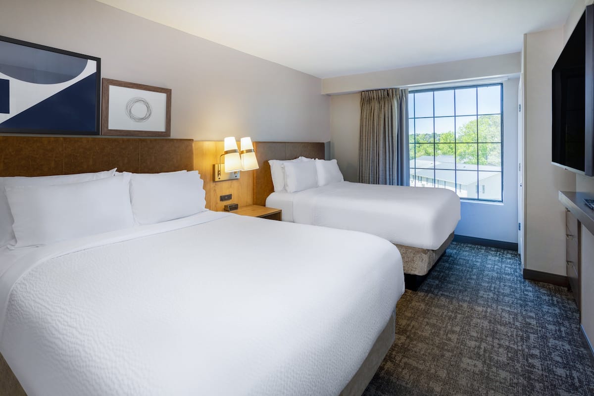 A hotel room with two beds, a large window, a flat-screen TV, and a wall-mounted artwork.