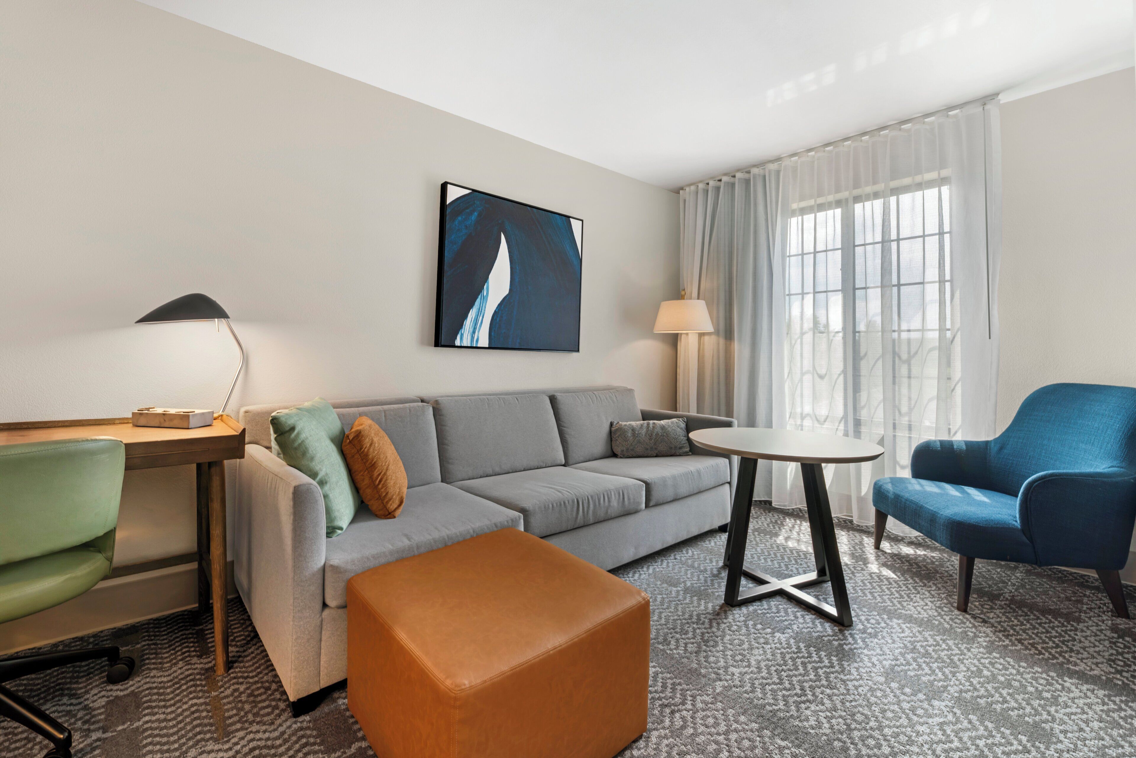 A modern hotel room with a grey sofa, a blue armchair, a small round table, and a desk with a lamp.
