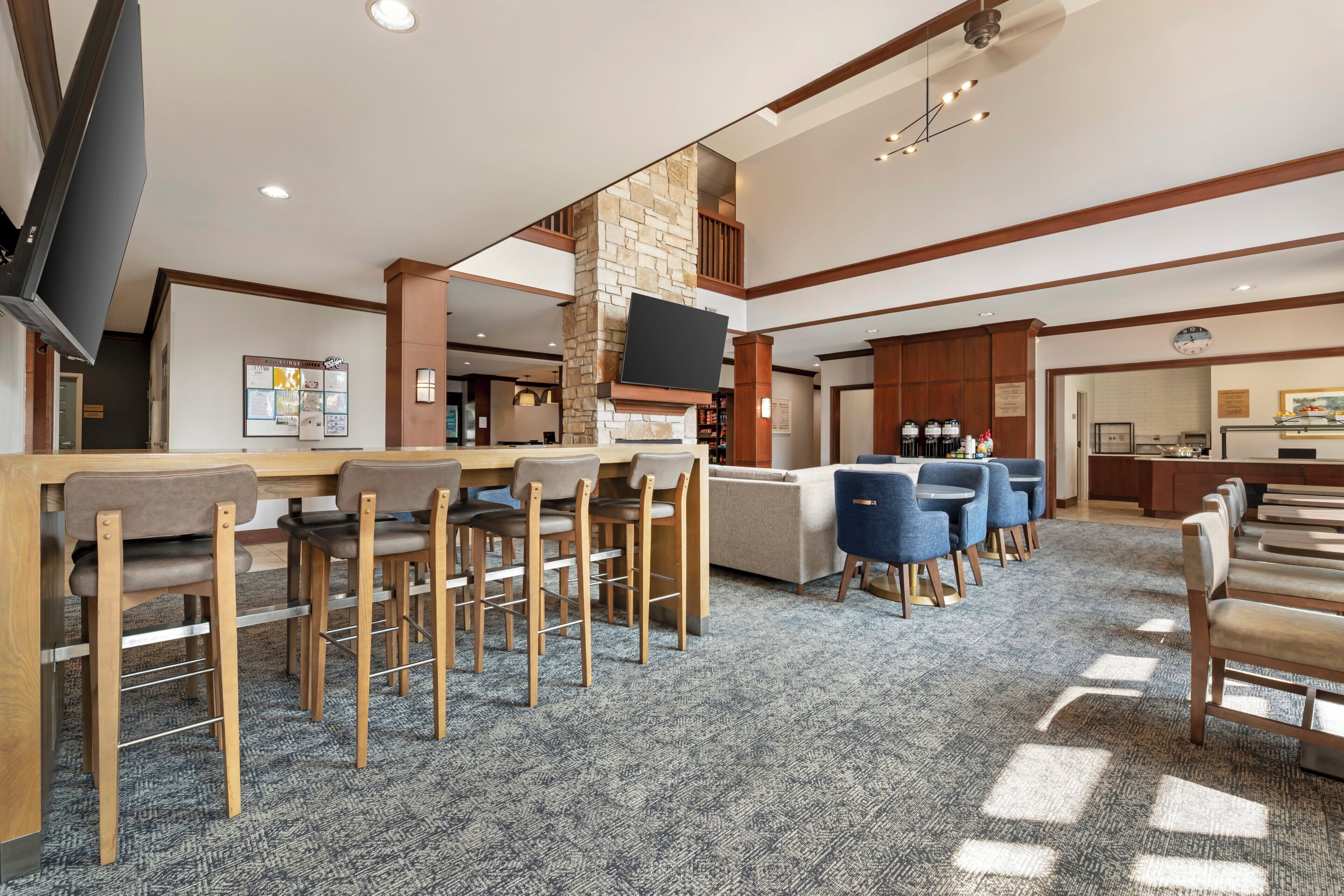 A hotel lobby with a bar area, seating, a television, and a kitchen in the background.