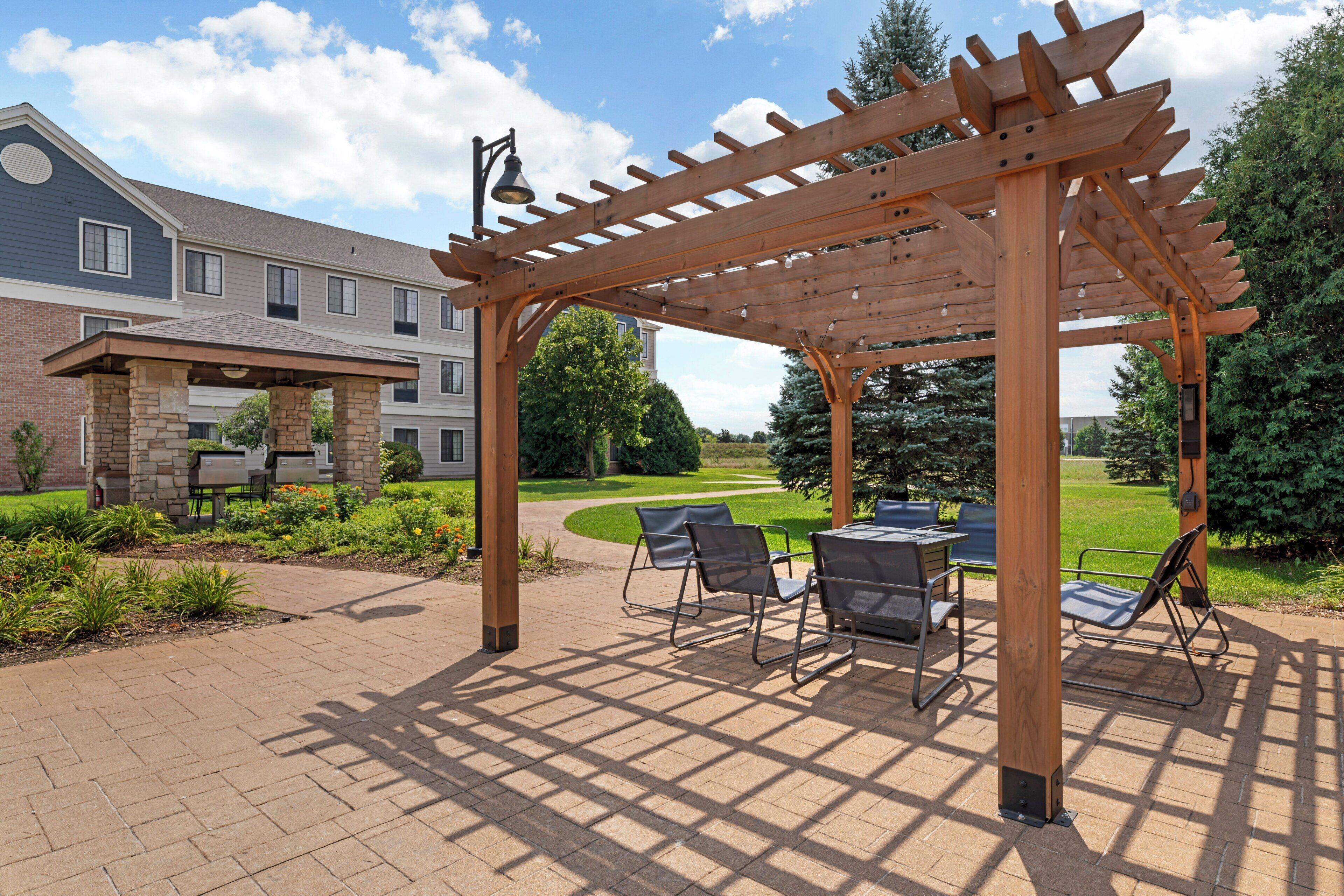 A wooden pergola with a table and chairs, a stone pillar, and a pathway leading to a building with a brick facade.