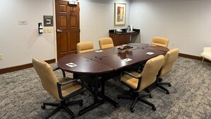 A conference room with a large oval table, six beige chairs, a wooden door, a small kitchenette, and a framed picture on the wall.