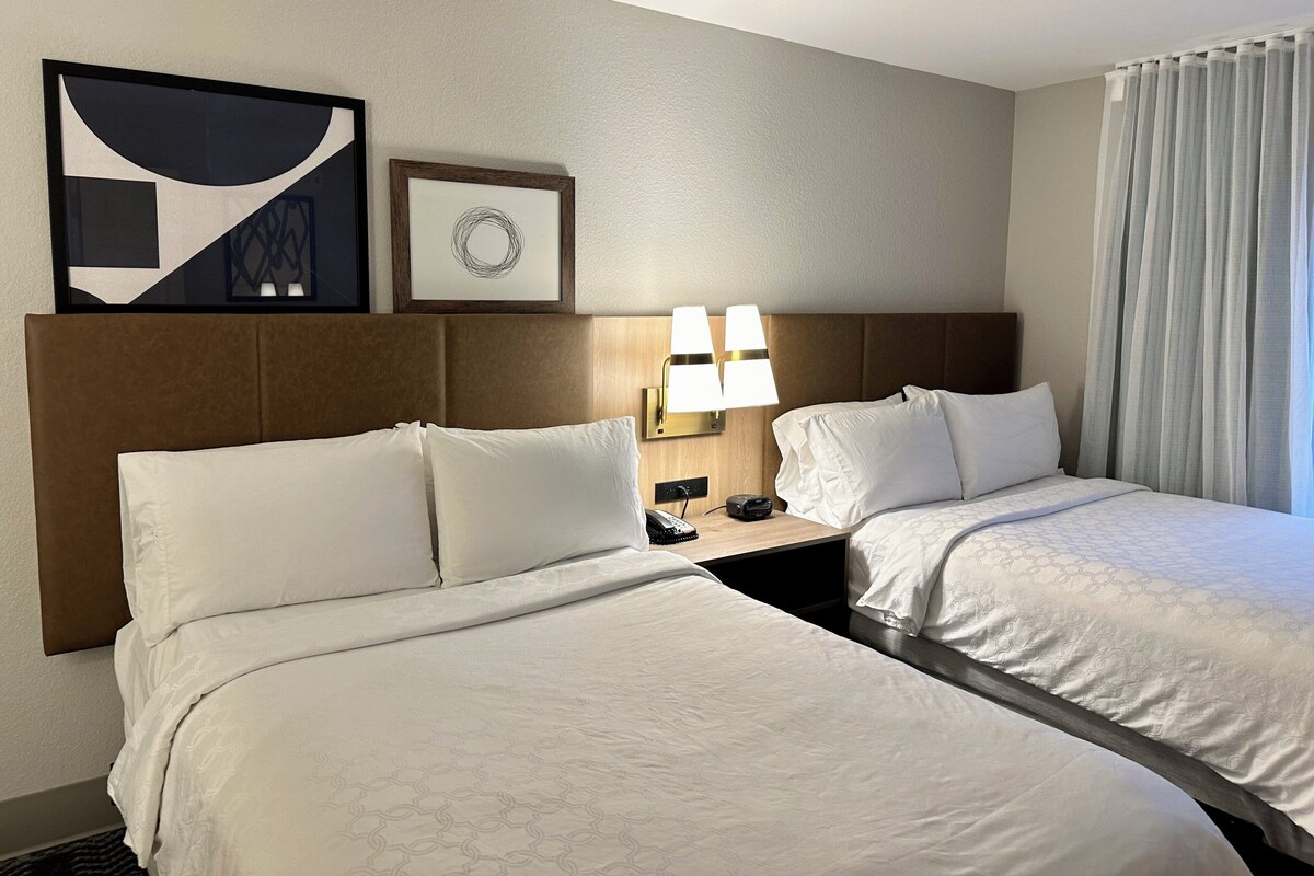 A hotel room with two beds, a headboard, a nightstand with a lamp, a phone, and artwork on the wall.