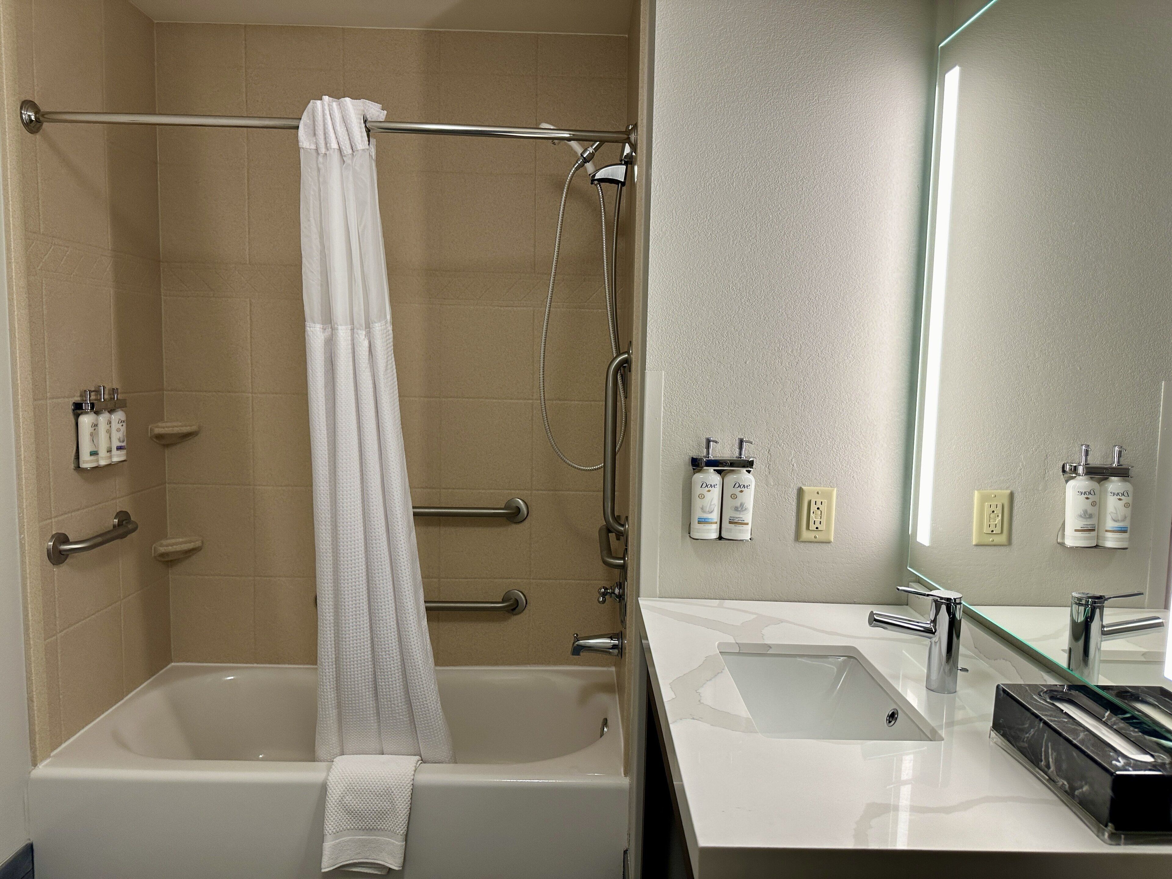 A modern bathroom with a bathtub, shower, and sink. There are handrails, a towel rack, and toiletries on the wall.