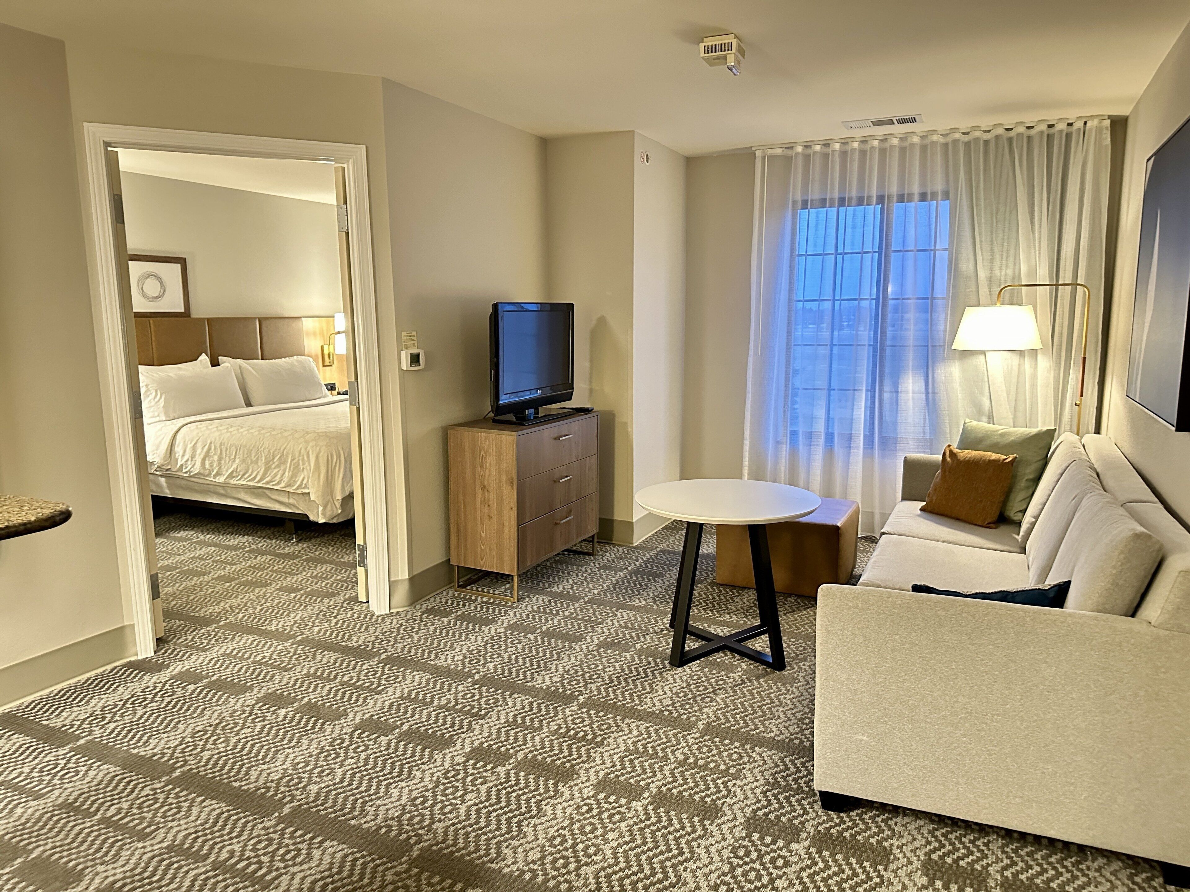 A hotel room with a bed, a TV on a wooden cabinet, a sofa, a small round table, and a window with curtains.