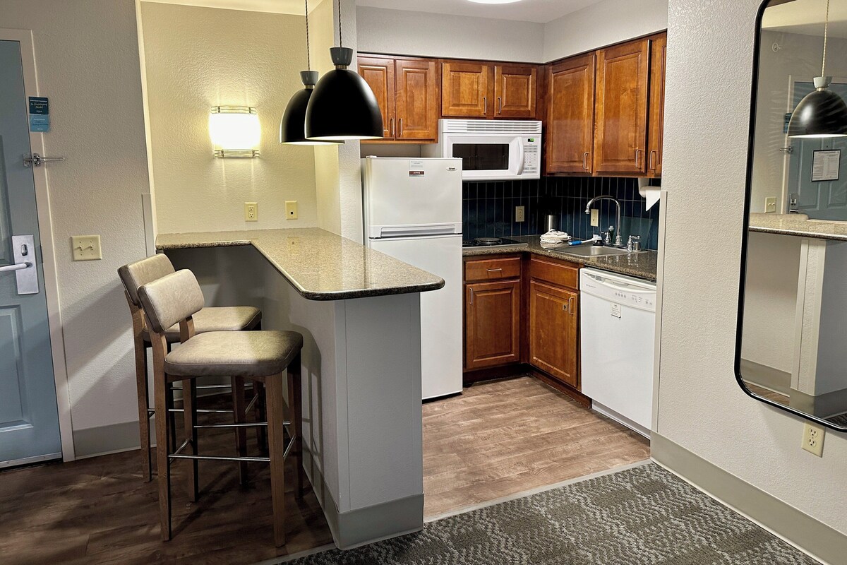 A compact kitchen with a refrigerator, microwave, and sink. There are wooden cabinets and a bar stool.