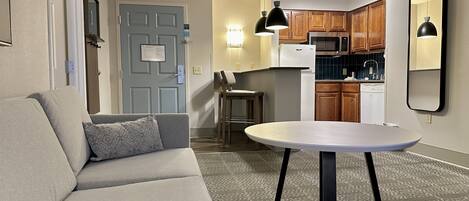 Un espacio habitable compacto con sofá, una pequeña mesa redonda, una kitchenette y una puerta con interruptor de luz.