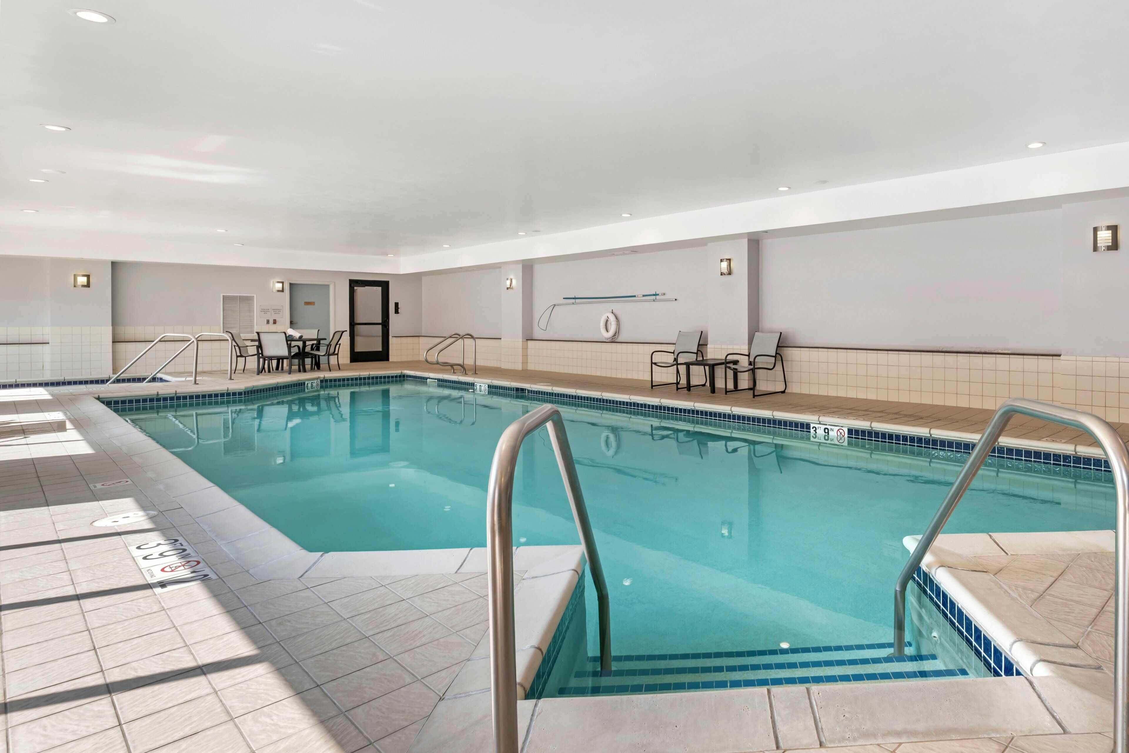 An indoor swimming pool with tiled walls, a door, and a seating area with tables and chairs.