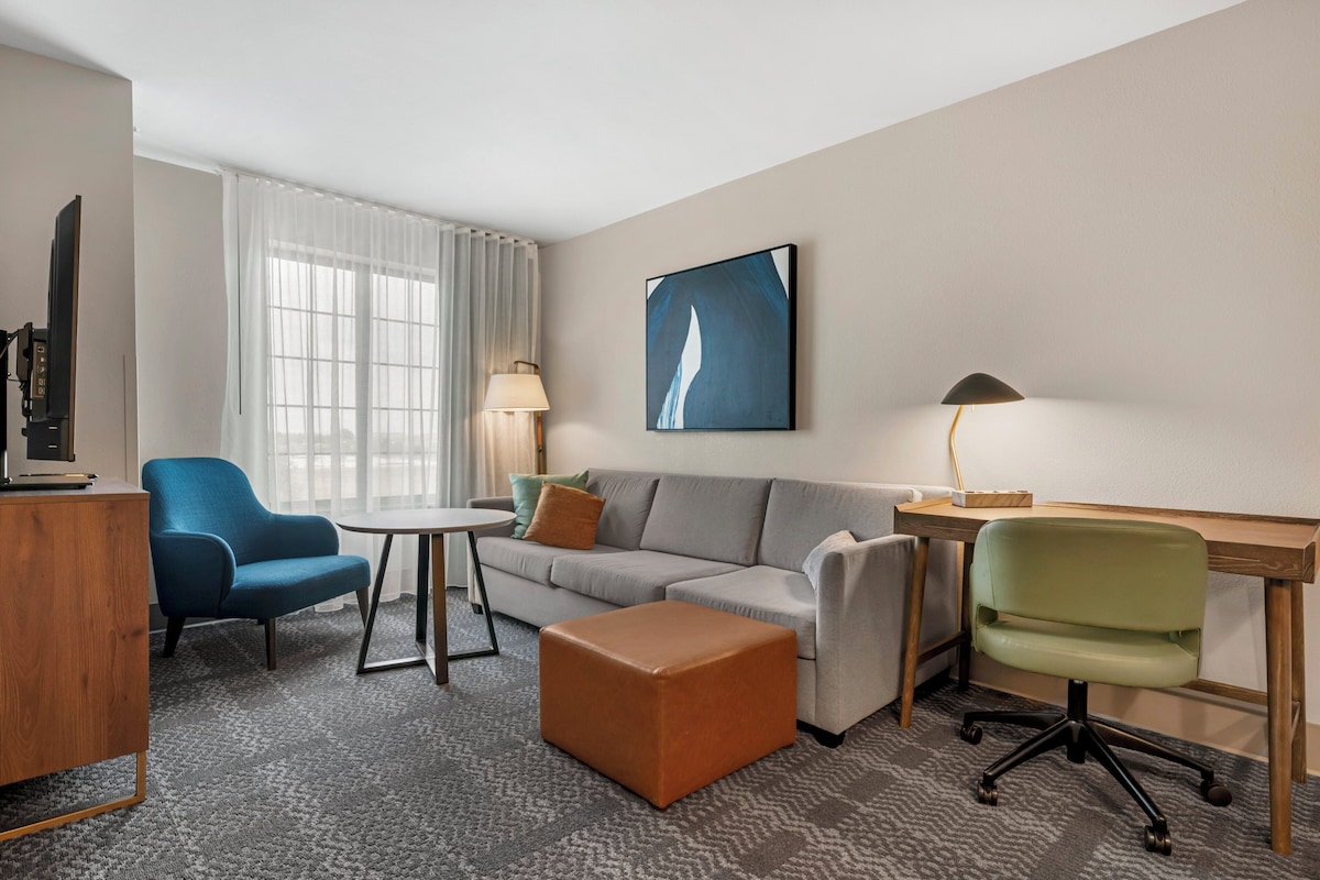 A modern hotel room with a grey sofa, a blue armchair, a wooden desk, a green office chair, a small round table, and a large abstract painting on the wall.