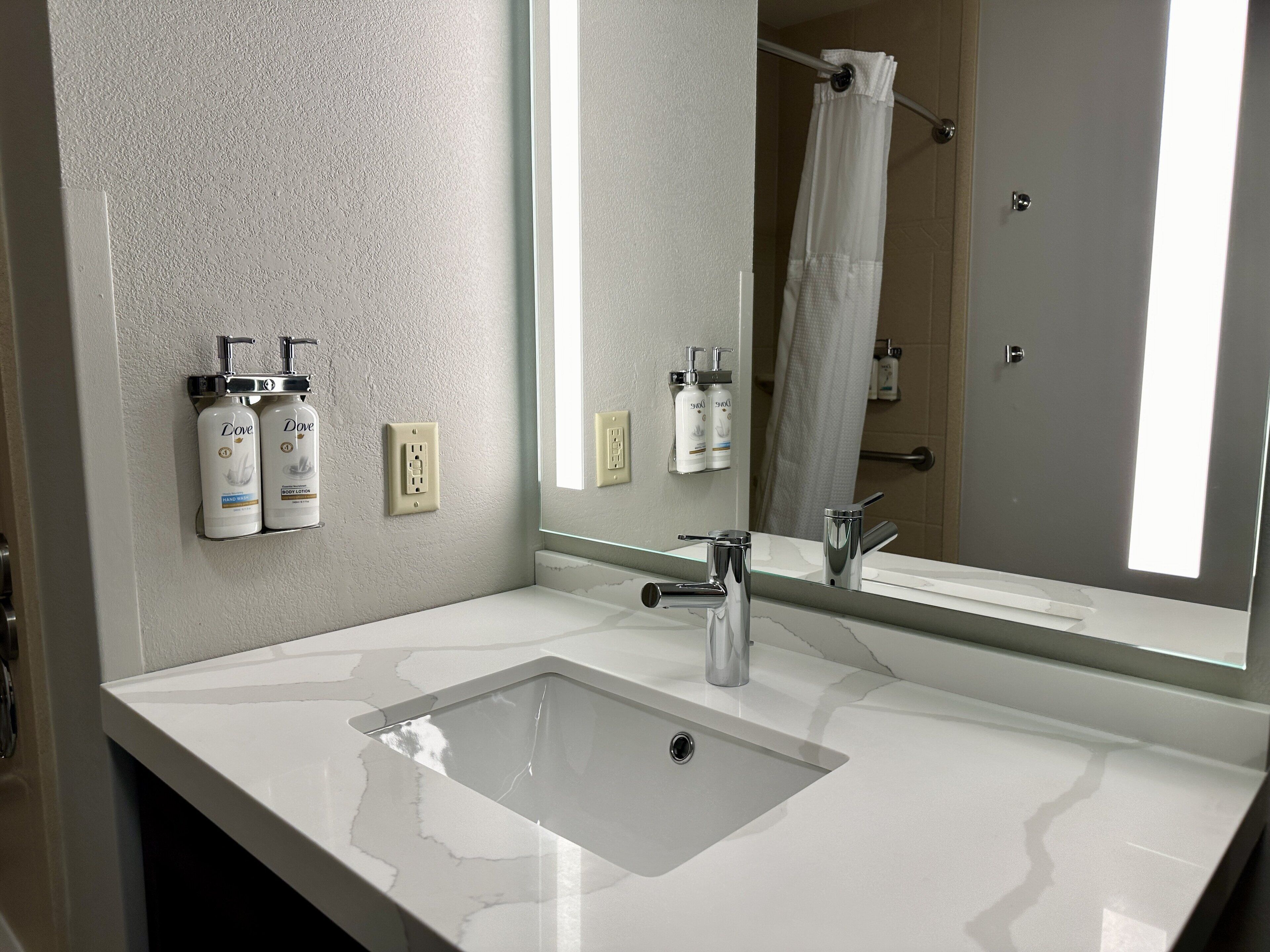 Un baño moderno con un lavamanos blanco, grifo cromado y un espejo grande. Hay dos botellas de jabón Dove fijadas en la pared.
