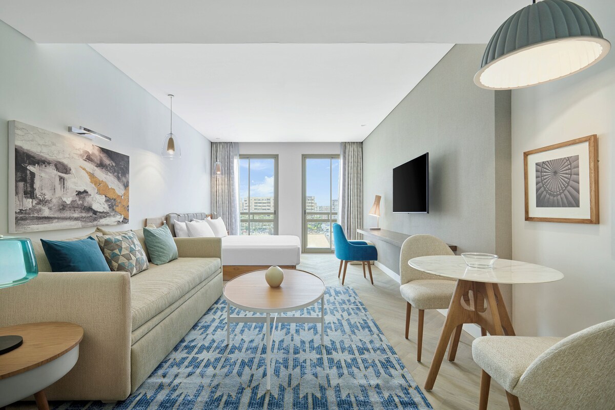 Habitación de hotel moderna con sofá, una mesita redonda, alfombra estampada azul, televisor y una ventana grande con vistas a la ciudad.