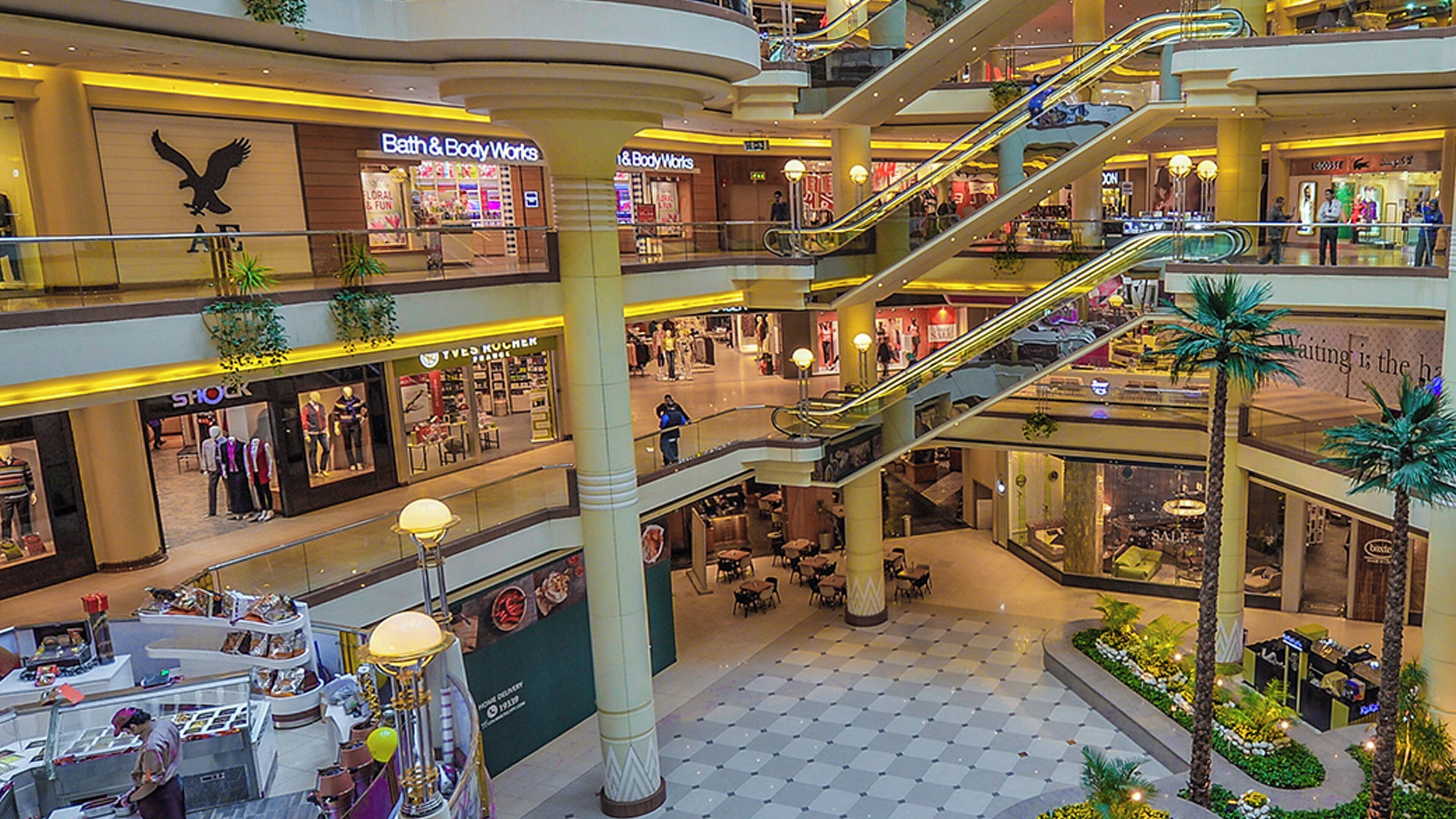 A multi-level shopping mall with escalators connecting floors, various retail stores, and a central open area with seating.