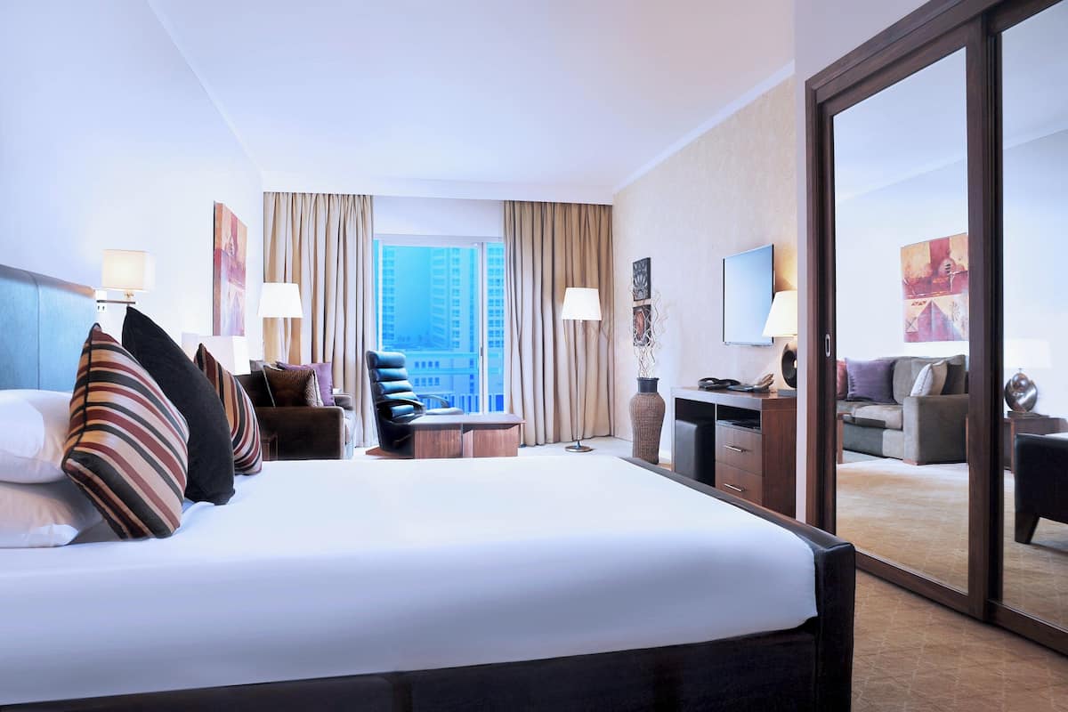 Quarto de hotel com uma cama grande, uma escrivaninha, uma cadeira, televisão e vista para edifícios altos.