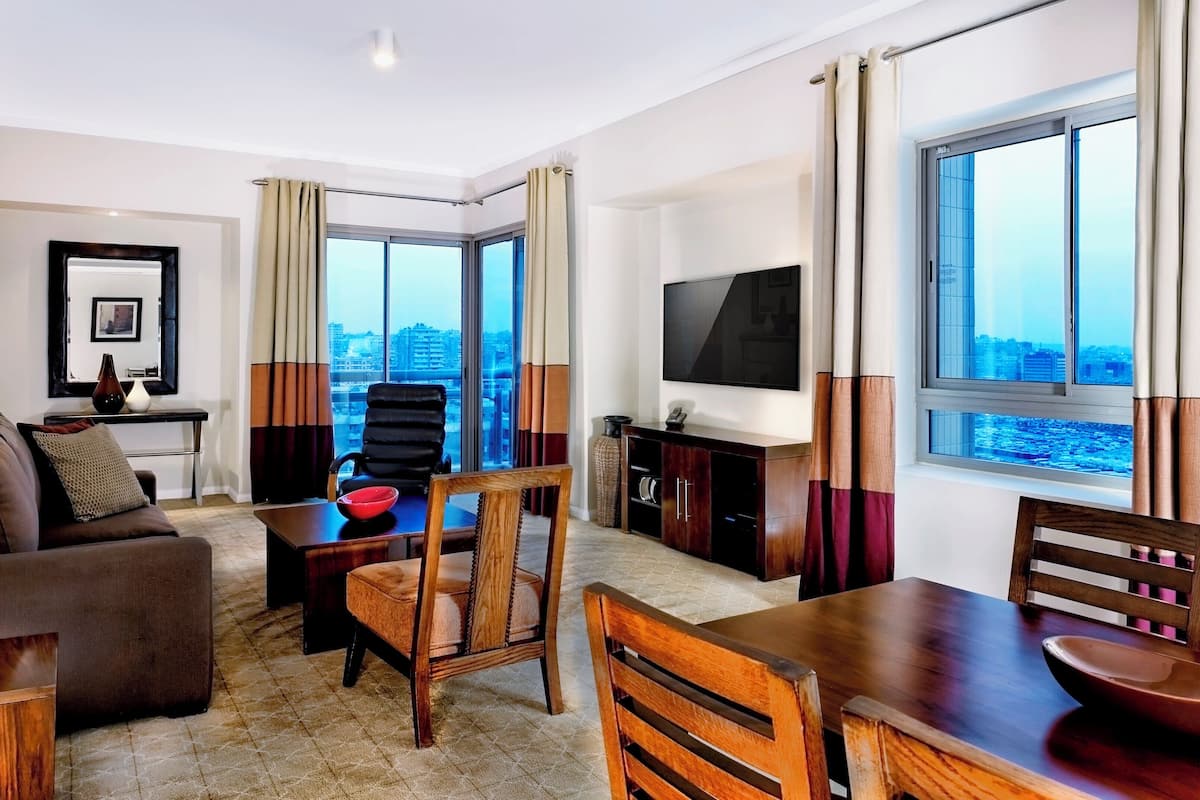 Quarto de hotel moderno com sofá, mesa de jantar, cadeiras, televisão e grandes janelas com vista para a cidade.
