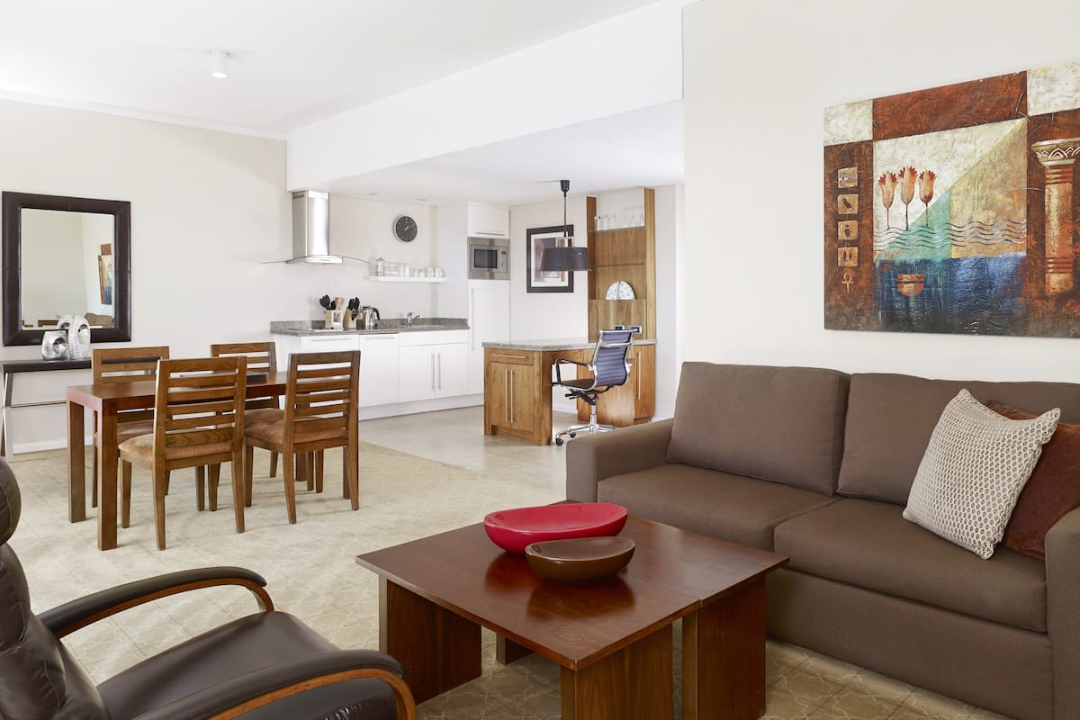 Uma sala de estar moderna com um sofá marrom, mesa de jantar de madeira e cadeiras. Uma cozinha visível ao fundo, com armários brancos e micro-ondas.