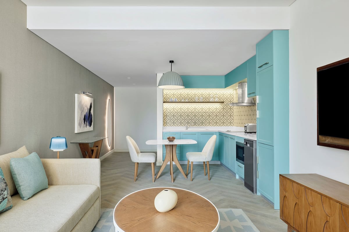 Uma sala de estar moderna com um sofá, uma mesa de centro e uma área de jantar com cadeiras e uma mesa. Uma cozinha com armários verde-água e bancada branca é visível ao fundo.