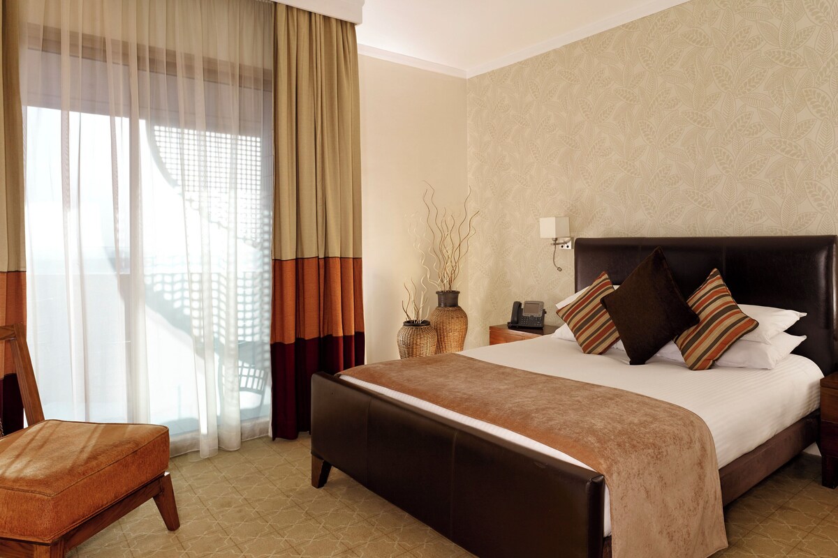 Una habitación de hotel con una cama grande, una silla, una mesita de noche y un ventanal con cortinas.