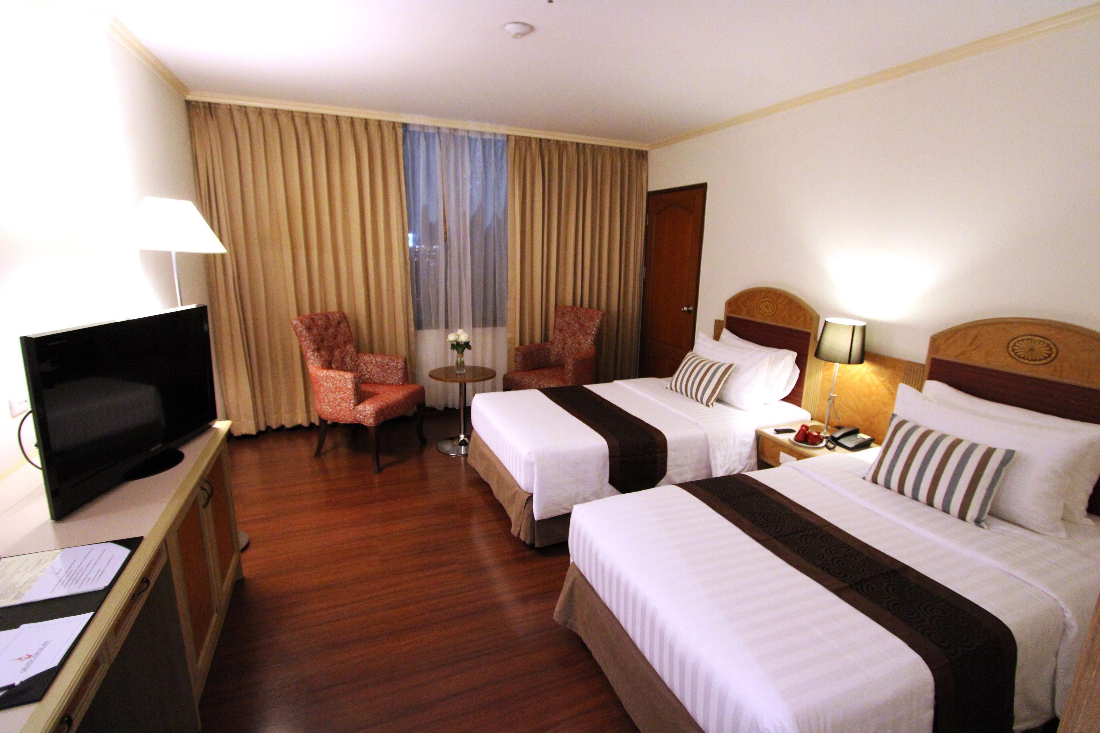 A hotel room with two beds, a television, a desk with a lamp, a chair, and a small table with a vase.