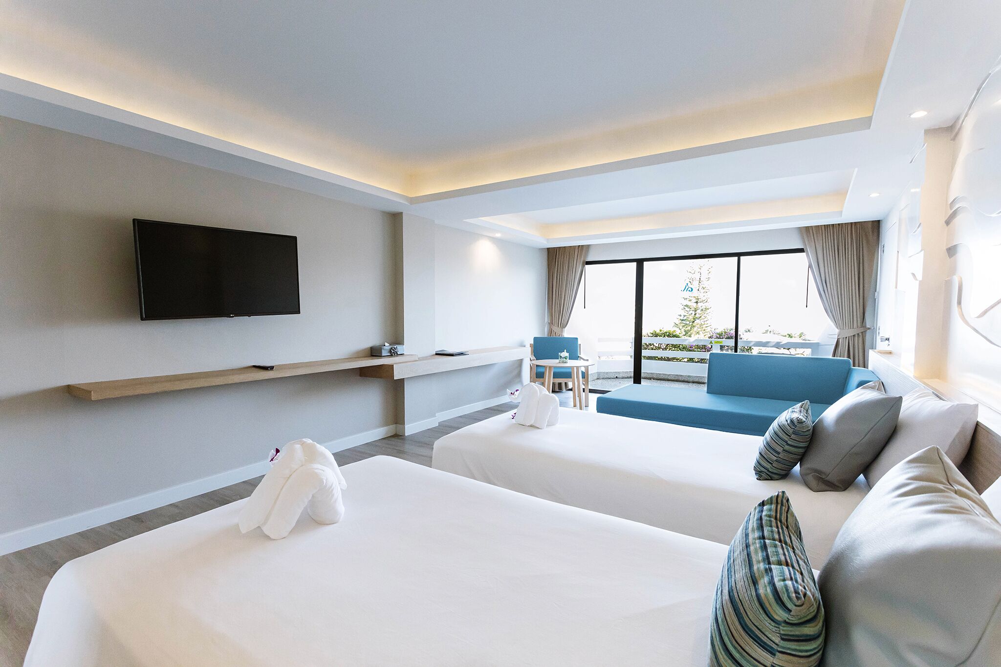 A hotel room with two beds, a TV mounted on the wall, a sofa, and a view of the outdoors.