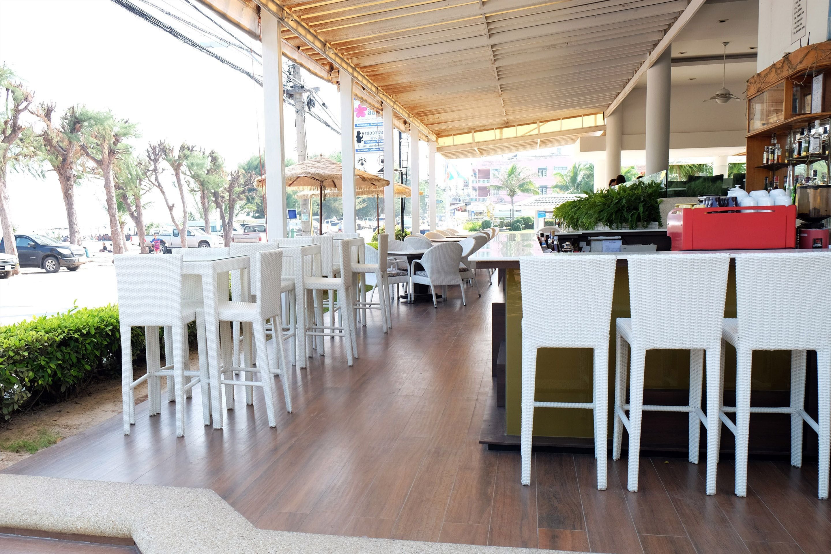 Ein Straßencafé mit weißen Korbmöbeln, einer roten Theke und Blick auf Palmen sowie geparkte Autos.