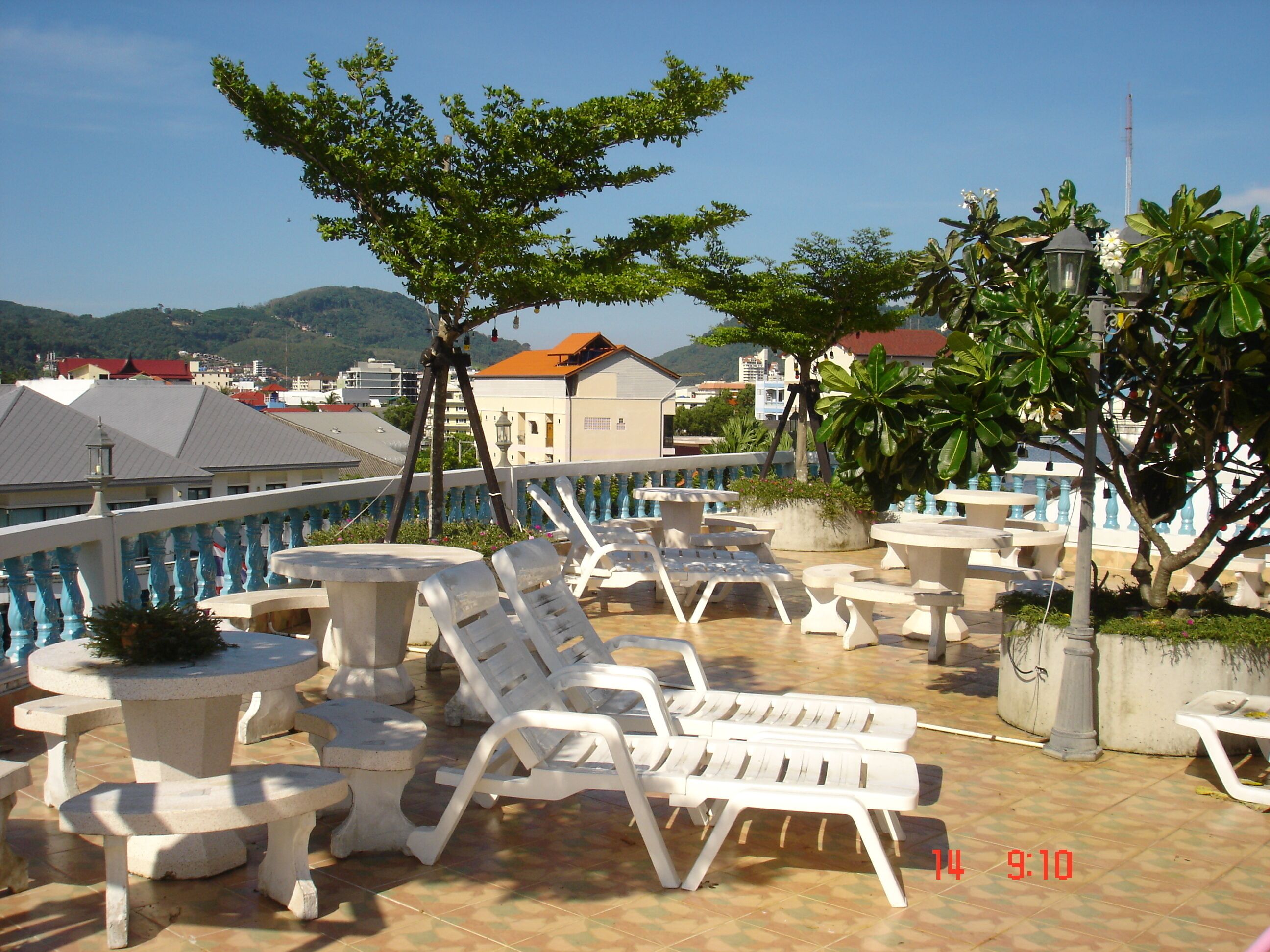 A terrace with white plastic chairs and tables, surrounded by greenery and overlooking a hilly landscape.