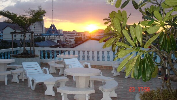 A terrace with white lounge chairs and tables, overlooking a sunset over a body of water with buildings in the background. - Lamai Apartment (Patong)